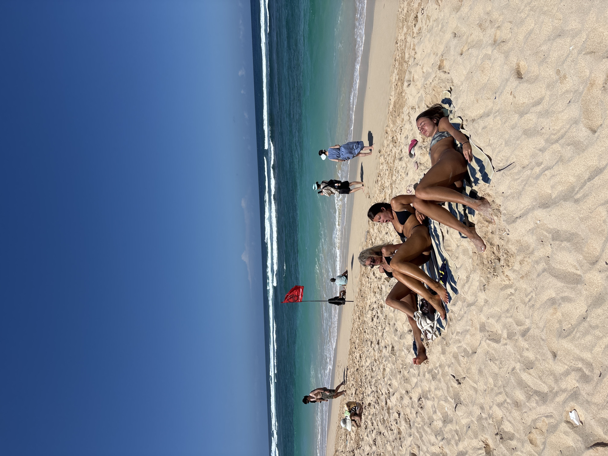 The image captures a sunny day at a beach in Kuta Selatan, Indonesia. The scene is dominated by a wide expanse of golden sand, with the turquoise ocean and clear blue sky in the background. The waves are gently rolling onto the shore.
In the foreground, three young women are sunbathing on towels. Two of them are lying on their backs, while the third is propped up on her elbows. They are all wearing bikinis.
Further back on the beach, several other people are visible. A man in shorts is walking along the shoreline, and a couple is sitting near the water. A red flag is planted in the sand, signaling caution for swimmers. The overall impression is one of a relaxed and leisurely day at the beach.