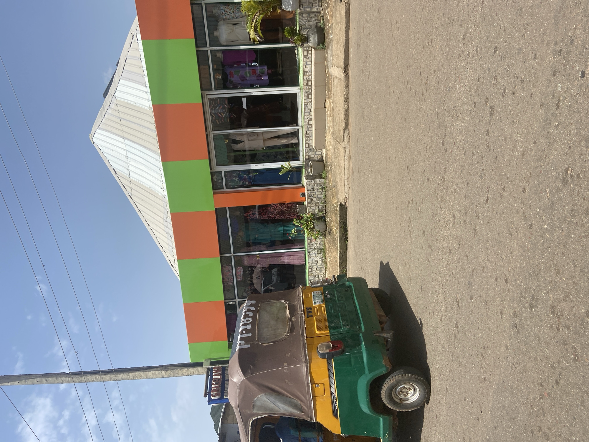 A three-wheeled auto-rickshaw, painted in yellow and green with a brown canvas top, is parked on an asphalt road in front of a shop. The shop has a striking facade with horizontal bands of orange and green. Large glass windows display mannequins dressed in clothing, suggesting it is a fashion boutique. The building has a peaked, corrugated metal roof. A utility pole stands to the left of the frame, with power lines crossing the blue sky. The background shows more buildings and greenery, indicative of an urban environment in Jos, Nigeria. The scene is illuminated by bright daylight, casting shadows on the road.
