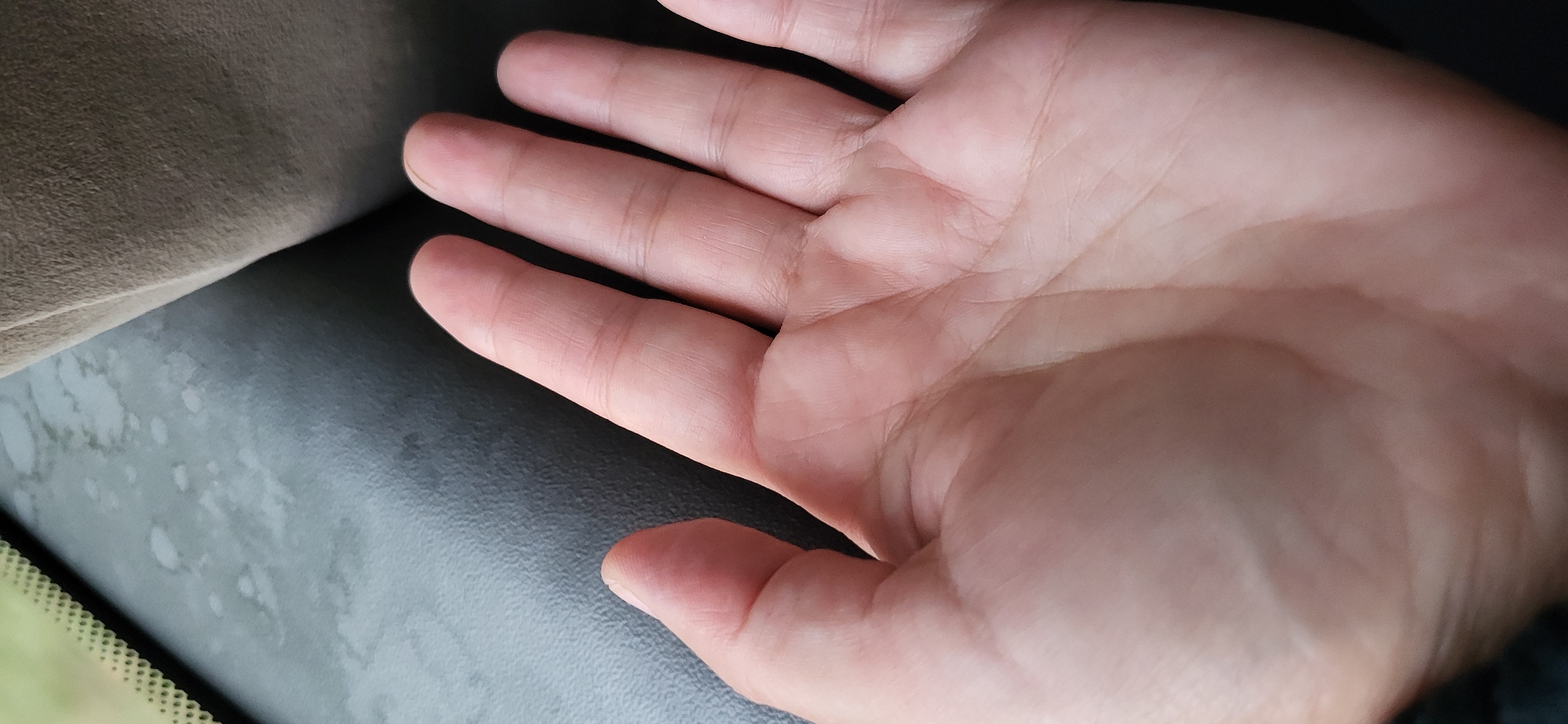 A close-up, eye-level shot focuses on the palm and fingers of a person's right hand. The hand is open, with the fingers slightly curled. The skin appears light-toned, with visible lines and creases on the palm and between the fingers. The background is out of focus, but suggests an indoor environment with textured surfaces. A portion of what appears to be a vehicle's dashboard or interior upholstery is visible in the upper portion of the frame, characterized by a dark, textured material and lighter, possibly water-stained, areas. The image was taken in Oskemen, Kazakhstan.
