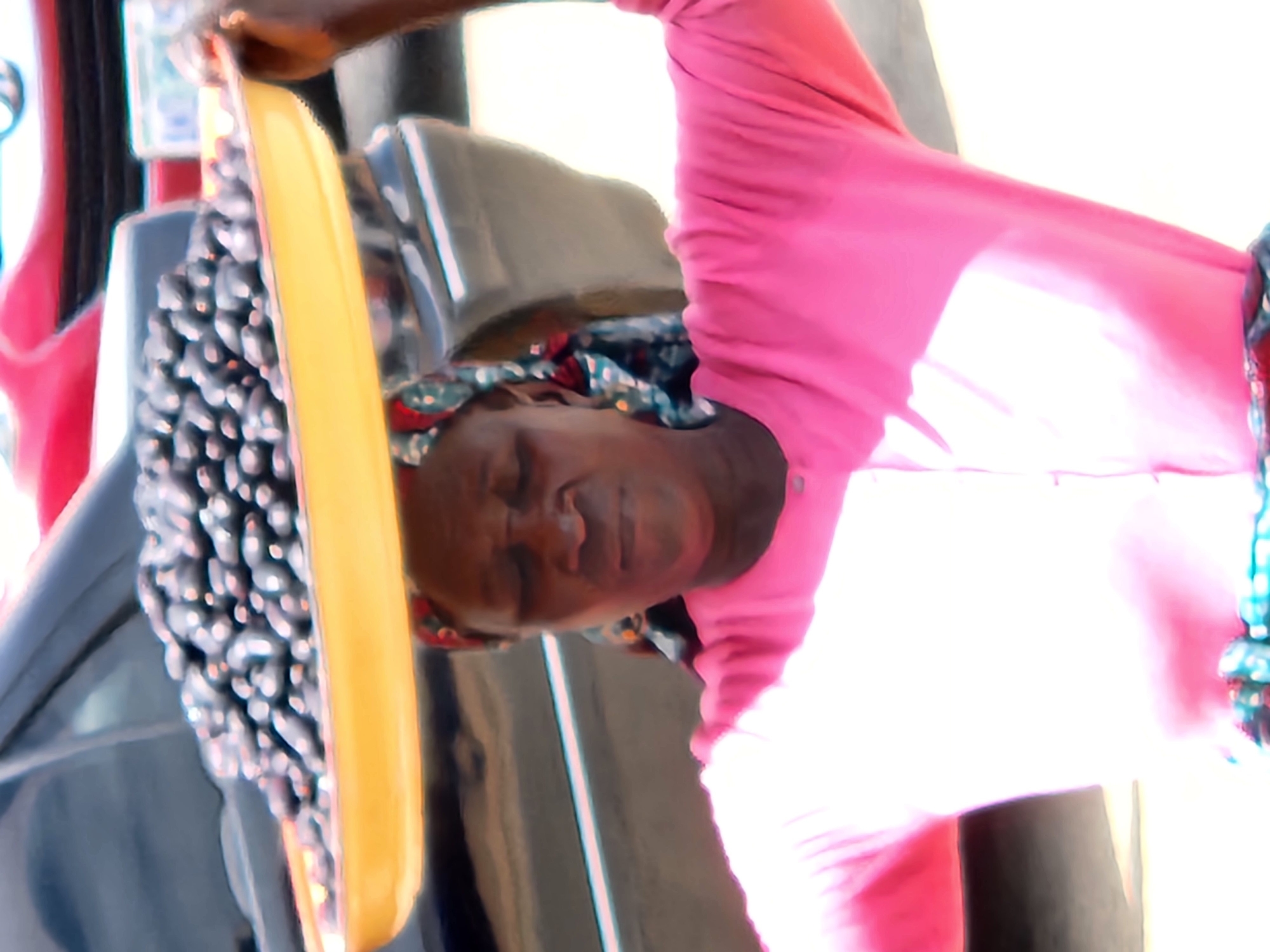 An outdoor medium shot captures a Black woman wearing a bright pink, long-sleeved shirt and a colorful headscarf. She is balancing a yellow tray filled with dark, oval-shaped fruits on her head. The fruits appear to be jamun or similar. Her eyes are closed, and her mouth is slightly open as she concentrates on maintaining her balance. Her arms are bent at the elbows and raised, with her hands supporting the tray from underneath. Behind her, a dark-colored car occupies much of the background, with hints of red and white vehicles visible further back. The scene suggests a marketplace or street setting in Jos, Nigeria, given the characteristic appearance of the woman and the types of goods often sold in such environments. The lighting indicates it is daytime, possibly midday, with bright sunlight casting shadows.