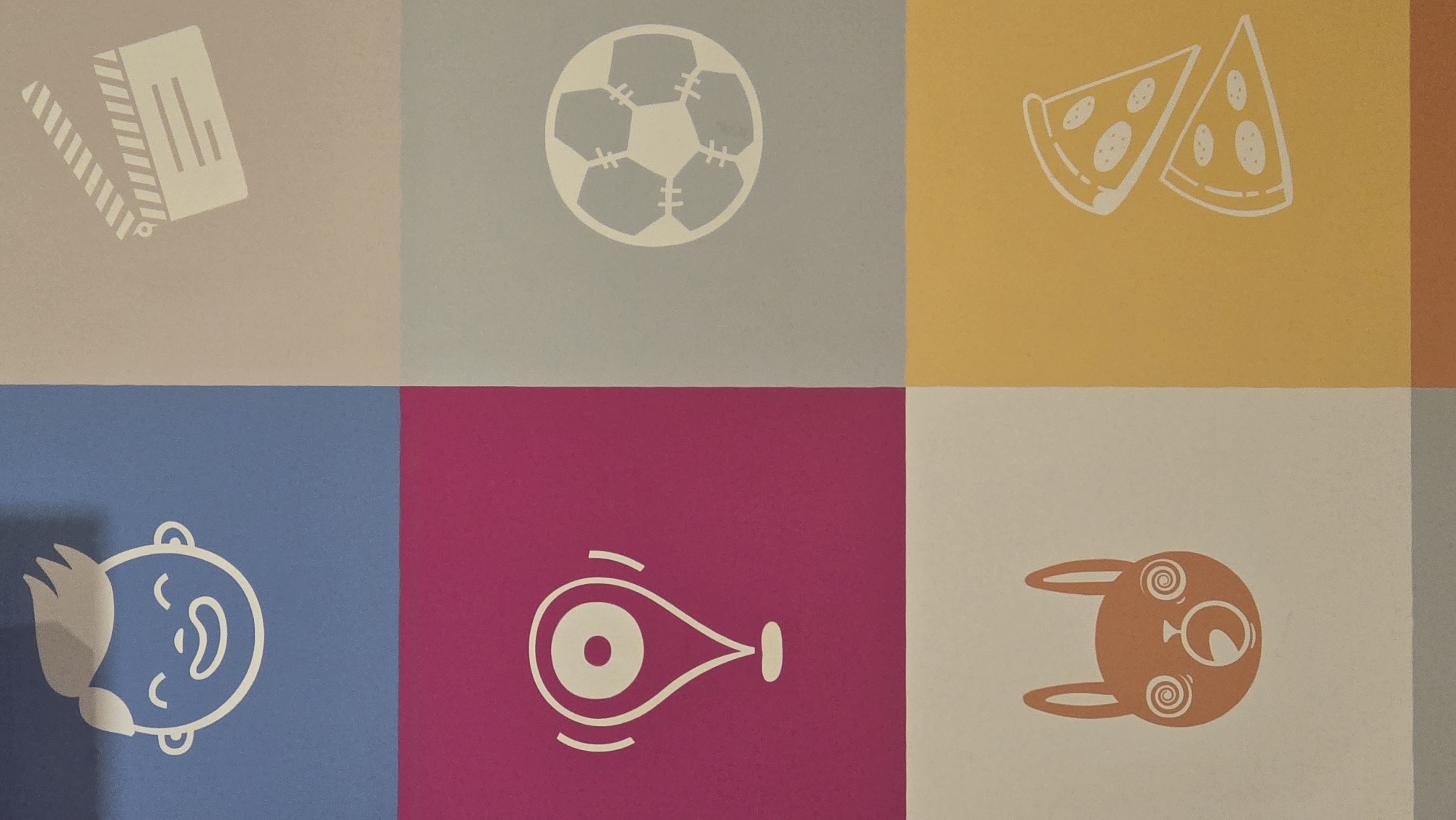 The image is a close-up, vertically oriented shot of a patterned wall. The wall is divided into six colored squares, with icons in the center of each. From top left, moving clockwise: a stylized white outline of a laughing baby's face on a blue background; a white outline of a film clapperboard on a light grey background; a white outline of a soccer ball on a medium grey background; two white slices of pizza on a yellow background; a white outline of a cartoonish, wide-eyed rabbit head with swirling pupils on a light grey background; and a white outline of a location pin icon on a magenta background. The image does not contain any information about the city of Chaikovsky, Russia.