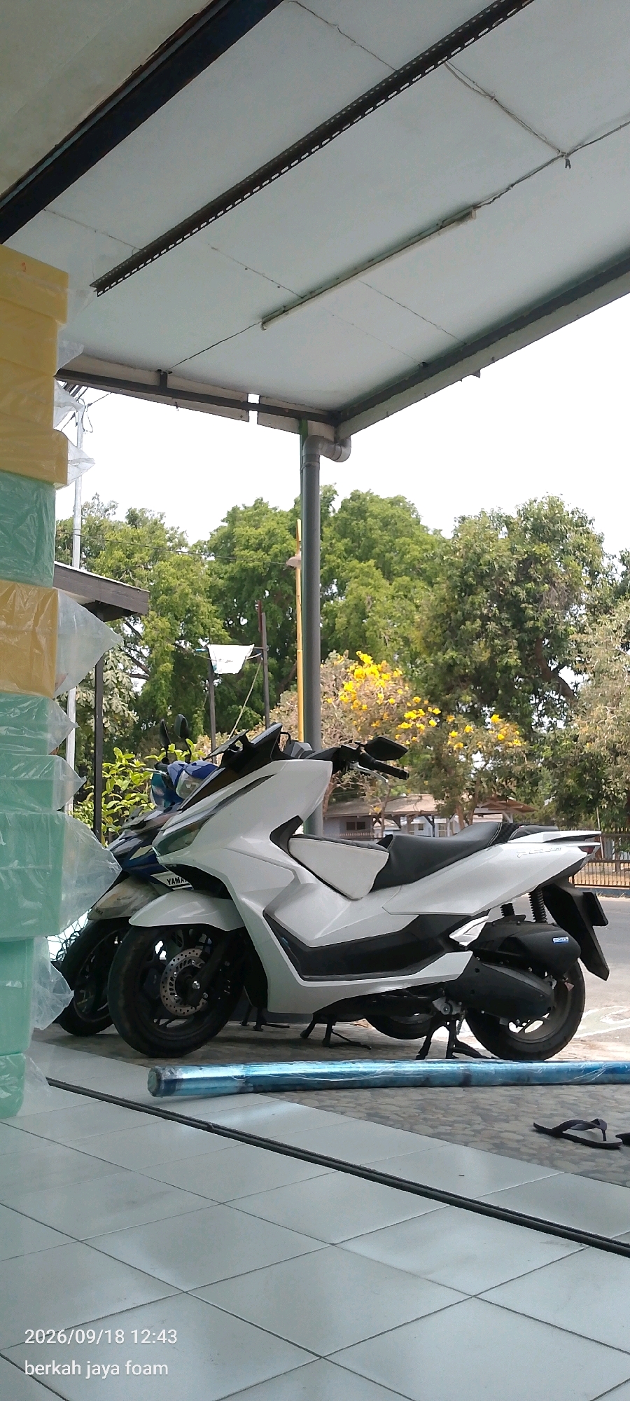 A white and black Yamaha scooter is parked on a tiled floor under a light-colored awning. To the left of the scooter, stacks of yellow and green foam wrapped in plastic are visible. Behind the scooter, another blue and white scooter is partially obscured. The background shows lush green trees and a yellow flowering tree, suggesting an outdoor setting. A thin, dark pole runs horizontally across the bottom of the frame, in front of a rolled-up blue item. The image is dated "2026/09/18 12:43" with the text "berkah jaya foam" below it. A pair of dark flip-flops are located on the tiled floor to the right. The scene appears to be in Kincang Wetan, Indonesia, possibly at a foam shop or a similar business.