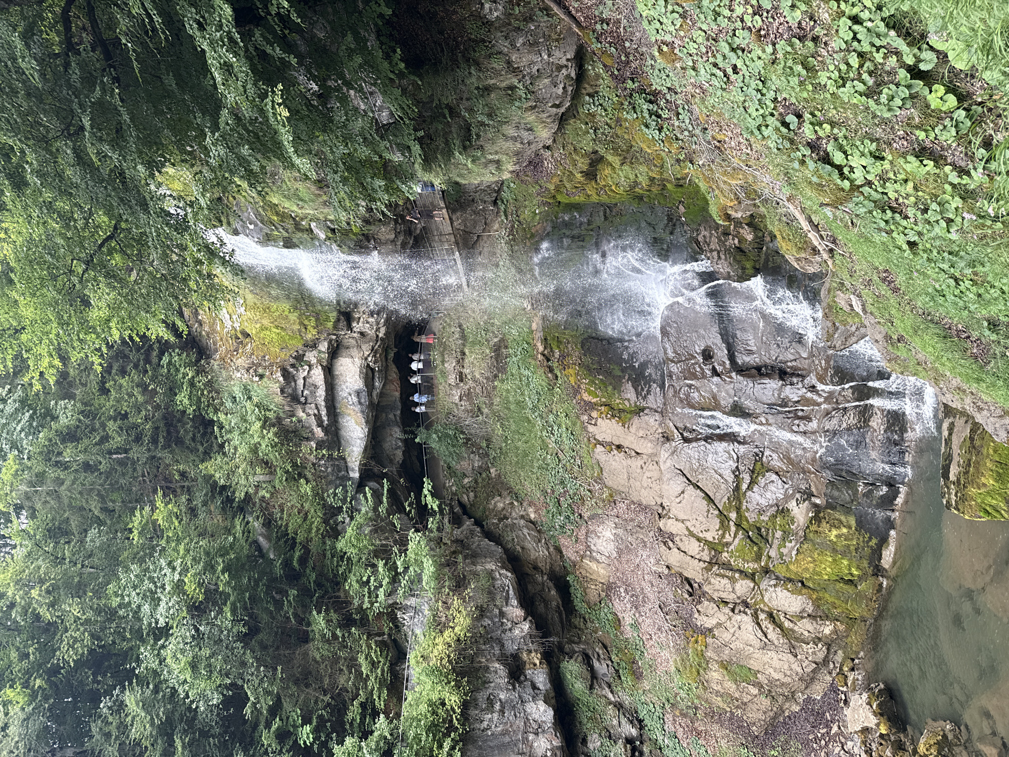The image shows a waterfall cascading down a rocky cliff face in Axalp, Switzerland. A narrow wooden walkway is constructed along the side of the cliff, allowing visitors to get close to the waterfall. Several people are visible on the walkway, some observing the waterfall and others walking along the path. The surrounding environment is lush and green, with dense trees and vegetation covering the cliffs. The water at the base of the waterfall collects in a clear pool. The overall scene depicts a natural landscape with elements of human infrastructure, creating a scenic view.