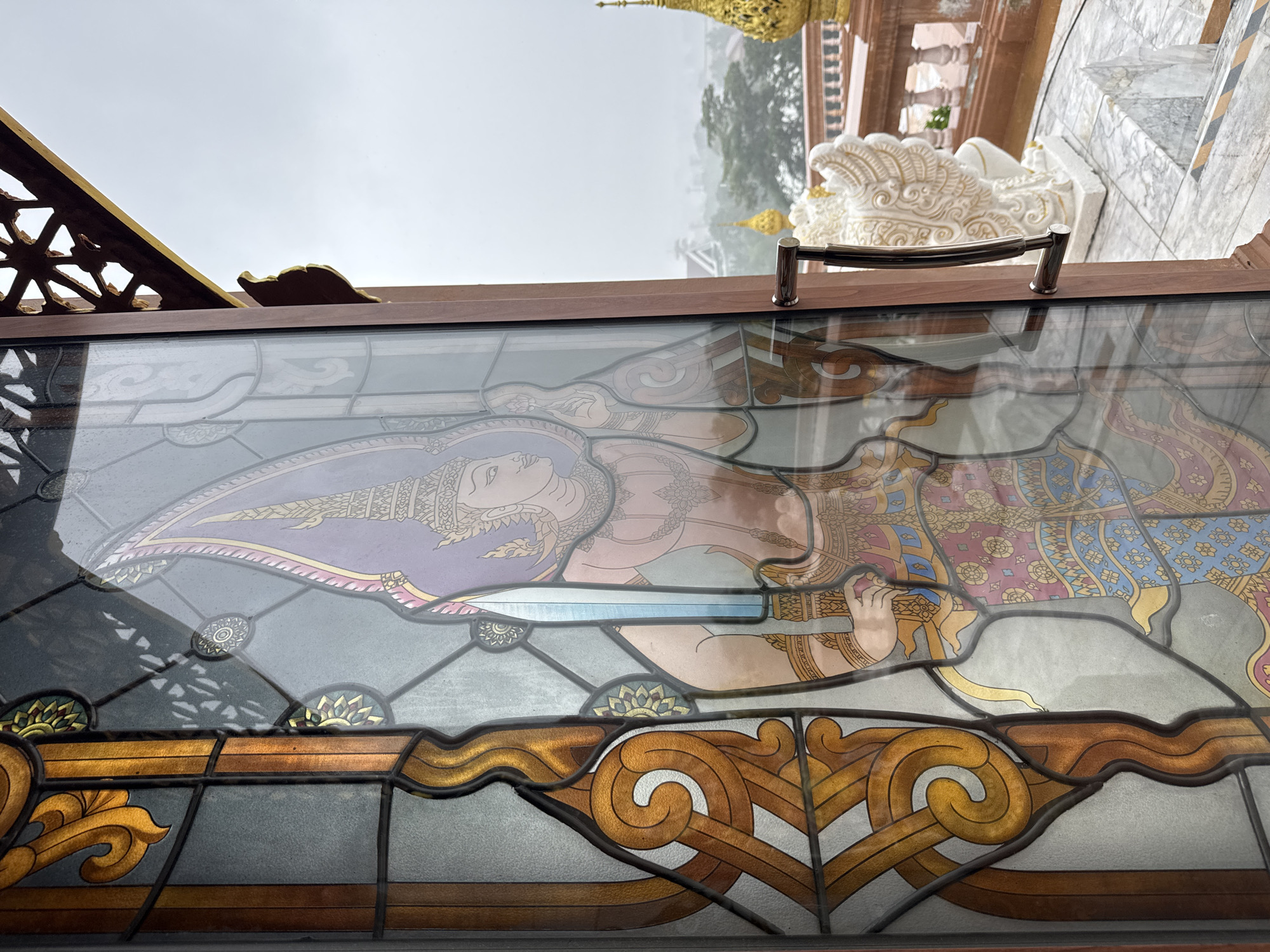 The image captures a detailed stained-glass window depicting a figure in traditional attire, holding a sword. The figure's head is adorned with an elaborate headdress, and their clothing features intricate patterns in pink, blue, and gold. The stained glass is framed by a wooden structure. Beyond the window, a hazy cityscape is visible, with glimpses of golden spires and ornate architectural elements, suggesting a temple or a similar religious or cultural site. A white mythical creature statue is also visible in the background. The overall ambiance suggests an overcast day. This scene is likely located at the Big Buddha Temple (Wat Phra Yai) in Phuket City Municipality, Thailand, given the characteristic architecture and iconography.