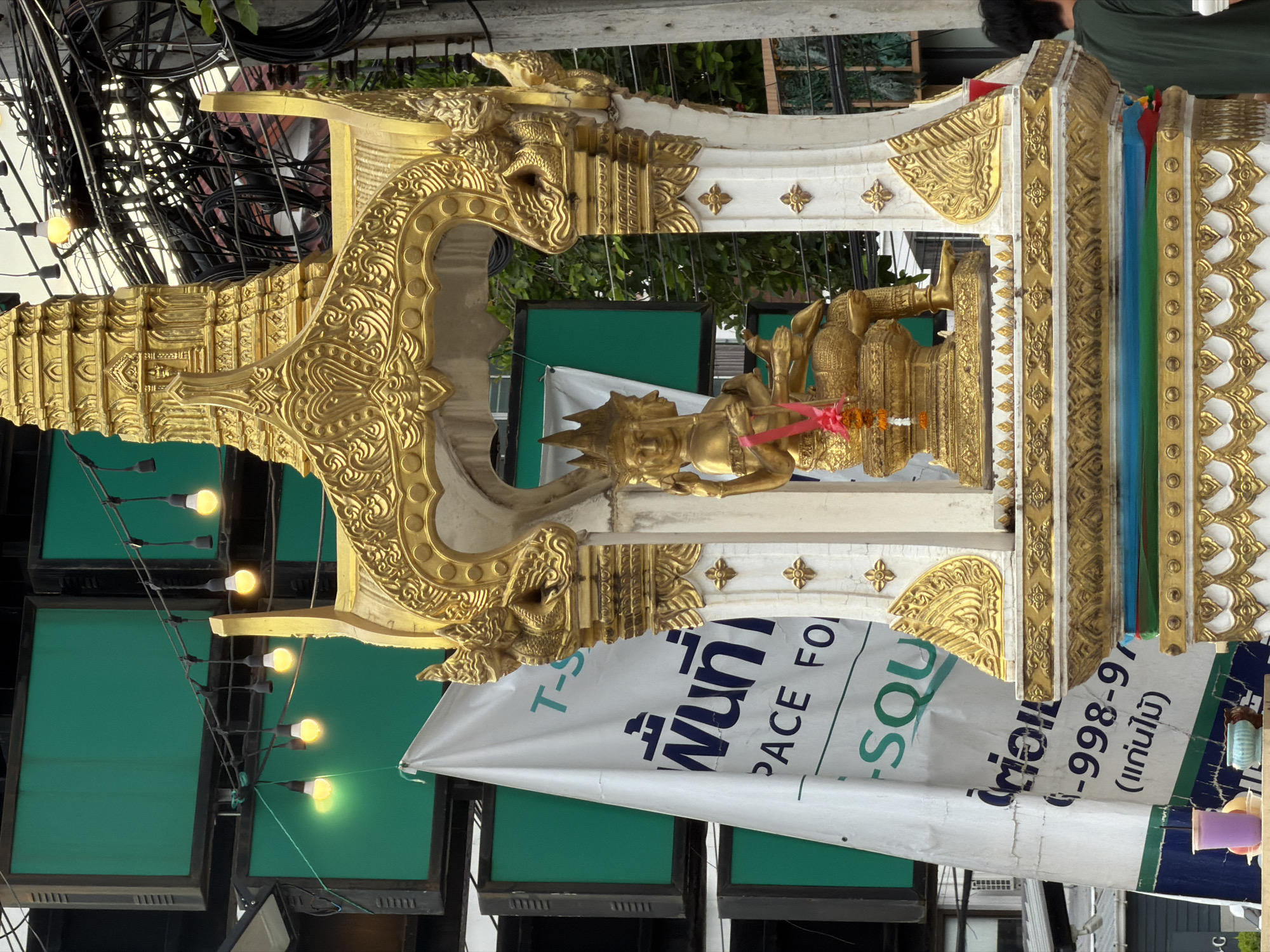 The image captures a vibrant street scene in Chiang Mai City Municipality, Thailand, focusing on a golden shrine adorned with intricate carvings. A multi-headed statue, likely a deity, sits at the center of the shrine, draped with a pink ribbon and flower garland. Behind the shrine, a large white banner with Thai script and English text, "SPACE FOR", is partially visible. The background reveals stacked green screens, electrical wires, and a glimpse of urban architecture. In the foreground, a person wearing a green shirt stands with their back to the camera, suggesting a casual urban setting. The overall impression is one of cultural richness and everyday life in a bustling Thai city.