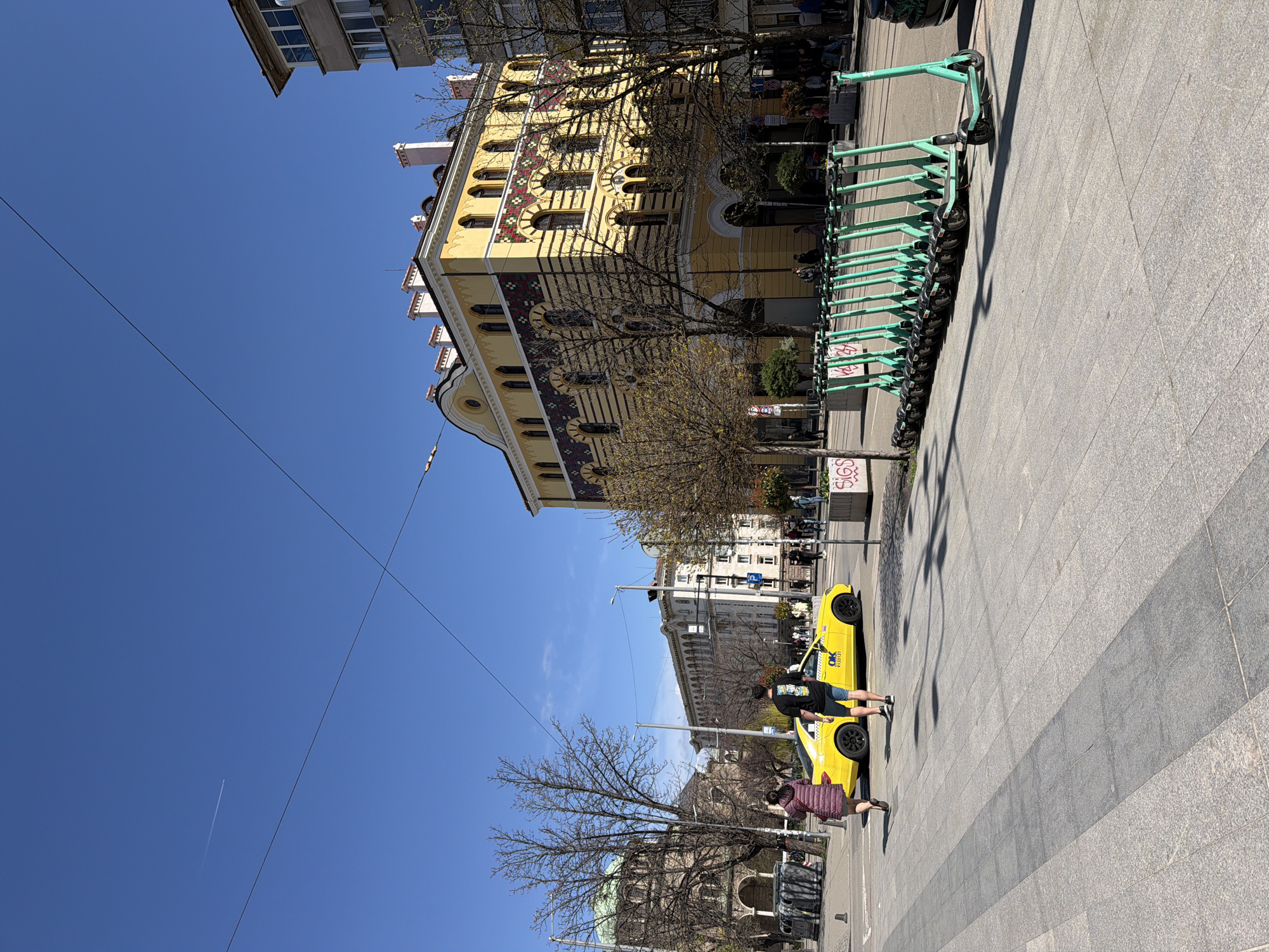 The image depicts a street scene in Sofia, Bulgaria, with a bright blue sky above. A distinctive yellow building with intricate mosaic patterns is visible on one side of the street, adjacent to a row of green electric scooters parked on the paved walkway. Two yellow taxi vehicles are parked at the roadside where two people are standing, one male and one female. Other buildings are seen further along the street. Bare tree branches are visible in the foreground.