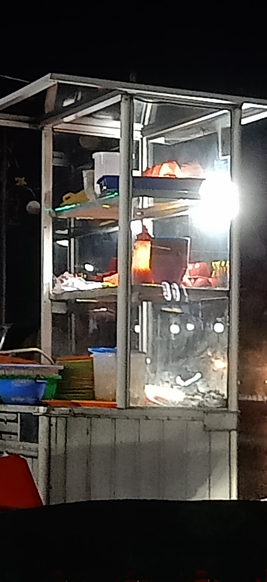The image depicts a brightly lit food stall at night, likely in Lhokseumawe, Indonesia, given the context. The stall is enclosed in a glass and metal structure, with multiple shelves displaying food items and containers. The lighting inside the stall is strong and somewhat glares on the glass, making it difficult to discern fine details of the food. However, some items appear to be in plastic containers and bottles, possibly condiments or ingredients. On the lower shelf, there are blue and green plastic tubs, suggesting preparation or storage of food items. The exterior of the stall is metallic and appears weathered. The background is dark, indicating it is nighttime, with some blurry lights suggesting other urban elements in the distance. The overall impression is of a bustling night market or a roadside eatery.