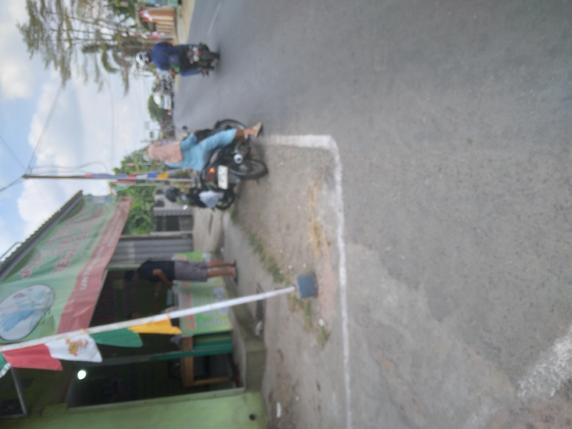 The image shows a street scene in Sokaraja, Indonesia. On the left, there is a green building with a banner that has text on it, but it is difficult to read due to the blurriness of the image. In front of the building, a person is standing near a table and appears to be interacting with someone inside the building. A flagpole with several flags is also visible in front of the building. On the right side of the image, there are two motorcycles on the road. One motorcycle has a person wearing a blue outfit and a headscarf riding on it. Another motorcycle further down the road has a person wearing a blue shirt and a helmet. The road appears to be paved, and there are trees and buildings visible in the background, suggesting an urban or semi-urban environment. The sky is partly cloudy.