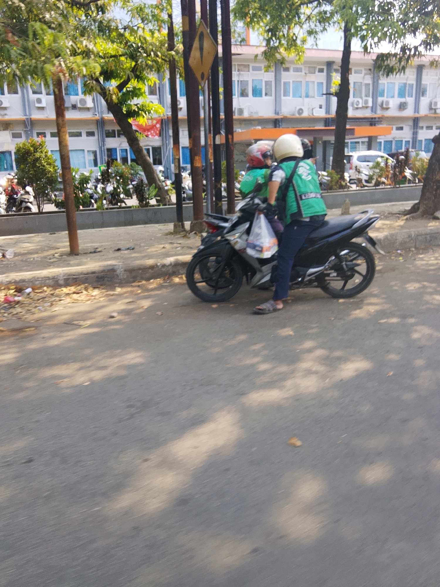 A motorcyclist wearing a green vest and a white helmet is seated on a black motorcycle, holding a plastic bag. The motorcycle is parked on the side of a street in Cirebon, Indonesia, with a building in the background. The building appears to be an office or institutional structure with multiple windows. Trees and other motorcycles are also visible in the scene. The ground is paved with asphalt and concrete. Sunlight casts shadows on the street.