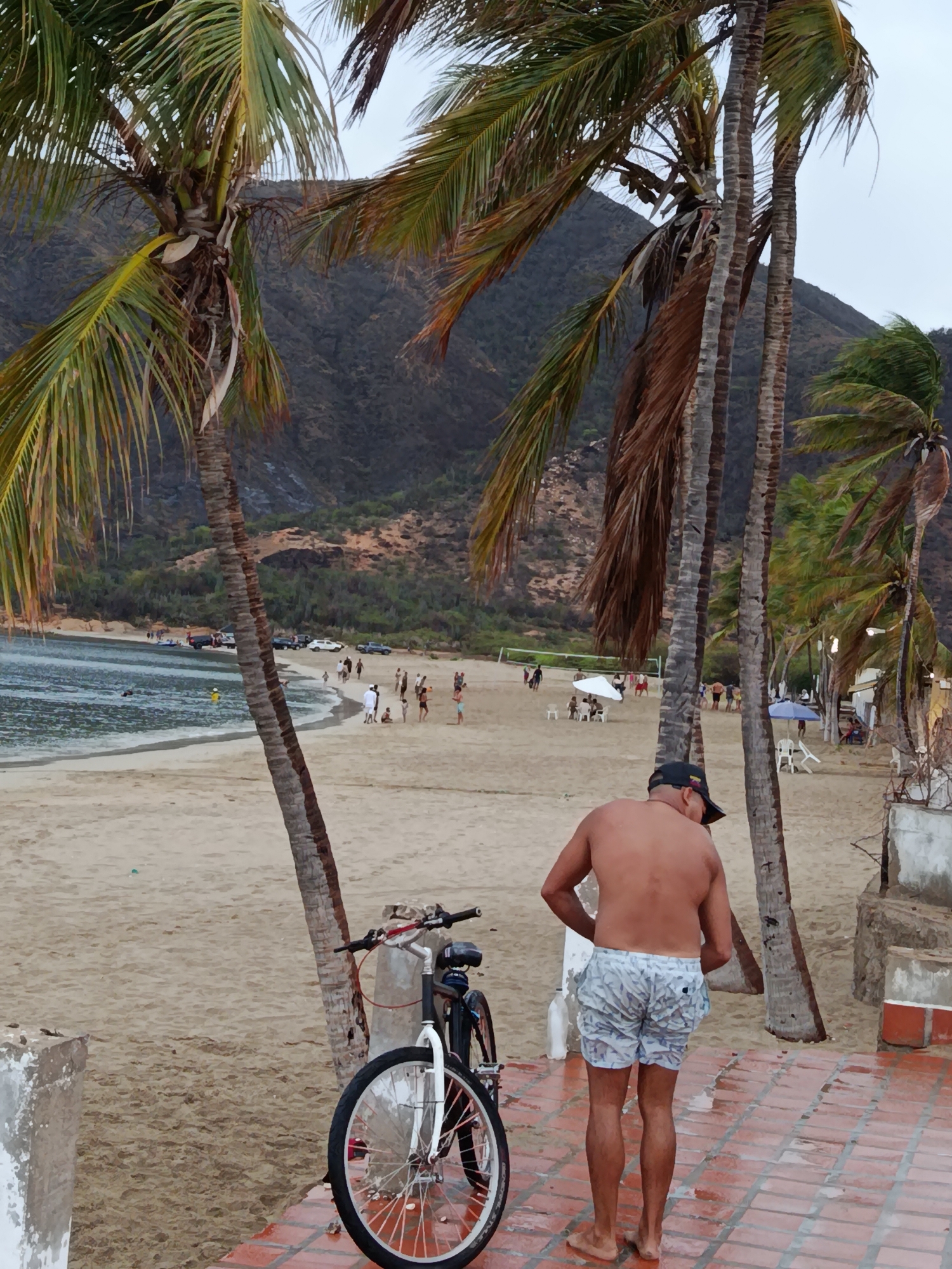 The image shows a beach scene in Juan Griego, Venezuela. In the foreground, a shirtless man wearing patterned shorts stands on a wet, tiled walkway, facing away from the camera. To his left, a bicycle with a white front wheel and black frame is parked. Beyond the walkway, a sandy beach stretches out, with palm trees framing the view. The ocean is visible to the left, with a few boats and people near the water. Further in the background, a crowd of people are gathered on the beach, some under umbrellas. A forested hillside rises behind the beach. The sky is overcast.