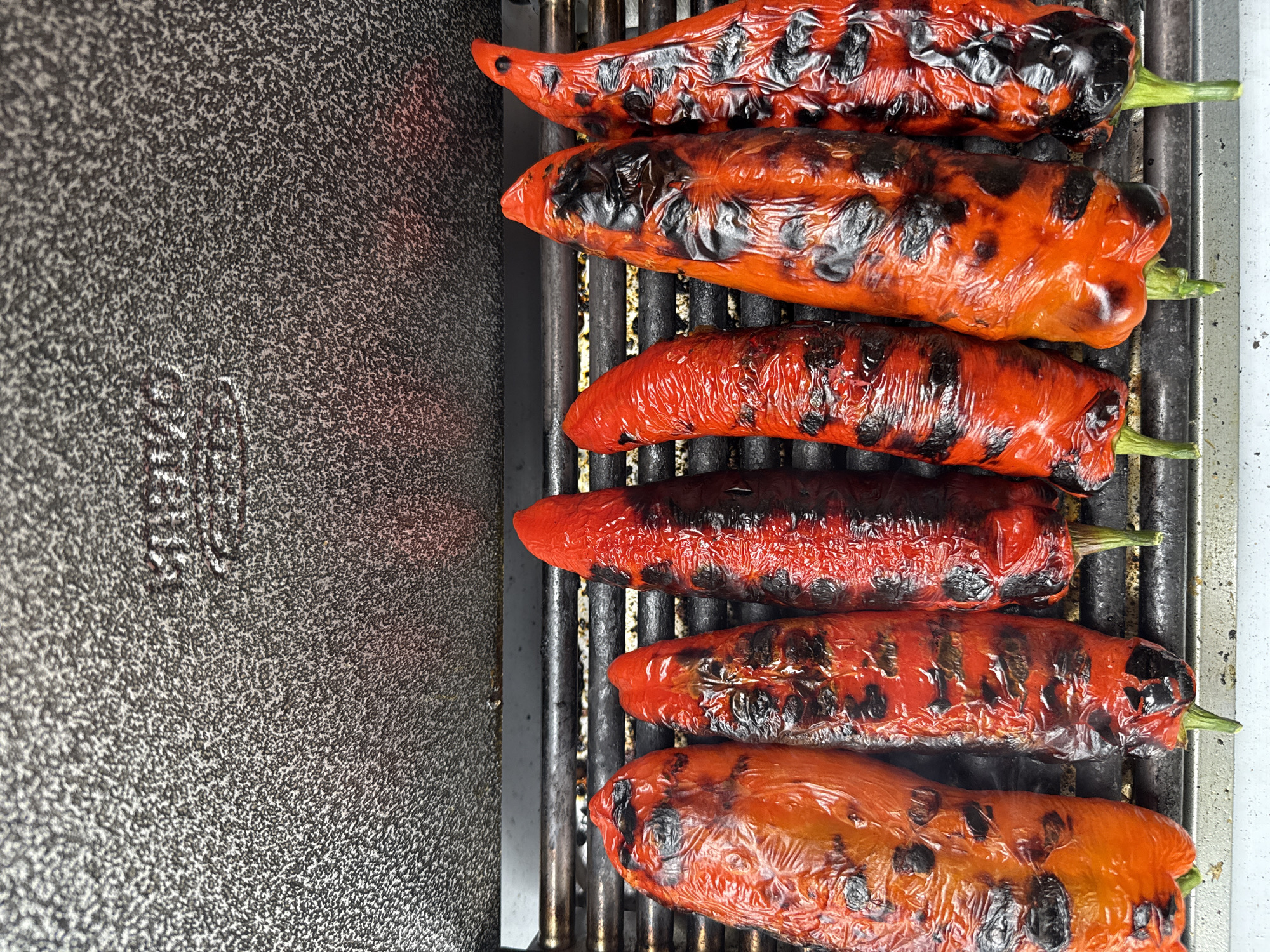 The image shows six red bell peppers being grilled on a metal grill. The peppers are charred in several places, with some areas appearing darker than others. The grill itself has metal grates and sits next to a textured grey surface. The scene doesn't have any recognizable London features. It appears to be a close-up photograph taken during the process of cooking the peppers.