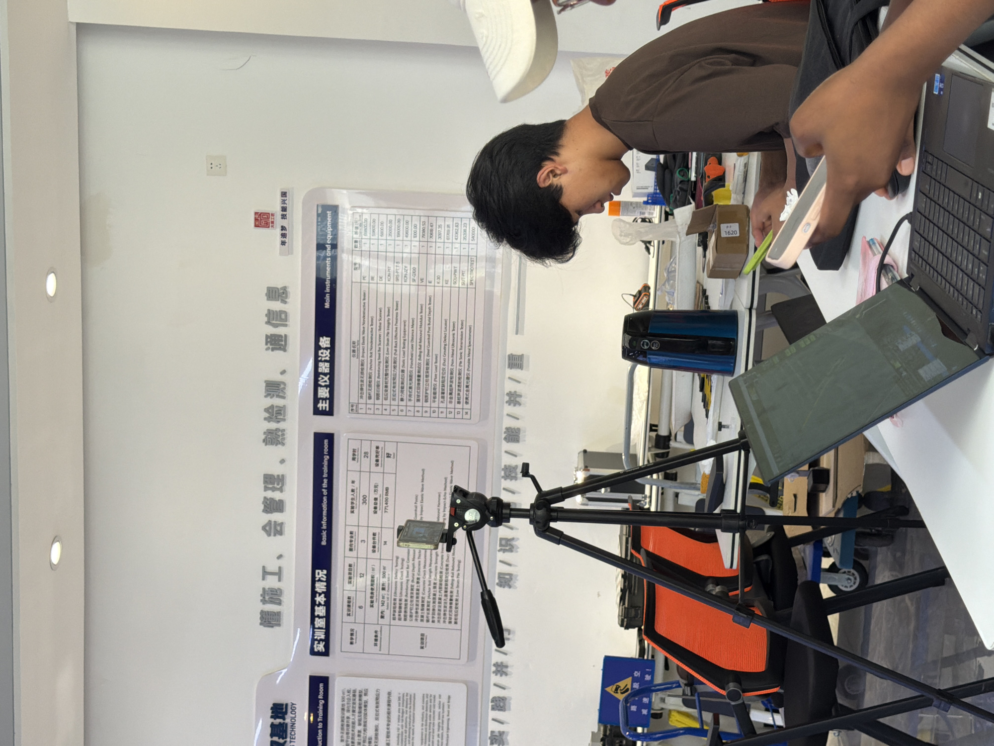 The photograph shows an indoor scene in a training room, possibly located in Jiangjin, China, indicated by the presence of Chinese text and what appears to be a company or institution logo with Chinese characters. The room is equipped with training materials and equipment. In the foreground, a young man is seated at a desk, looking down at a smartphone in his hand. Another person's hand is visible holding a phone, also likely engaged with a device. A laptop is open on the desk in front of the person holding the phone.
The background features a large informational poster with charts and text in both Chinese and English, detailing "Basic Information of the training room" and "Main instruments and equipment." A tripod with a camera is set up in the middle ground. Various pieces of equipment and furniture, including chairs and carts, are visible in the room, suggesting a functional training or workshop environment. The lighting appears to be artificial, with a ceiling light fixture visible. The overall impression is that of a contemporary educational or vocational training facility.