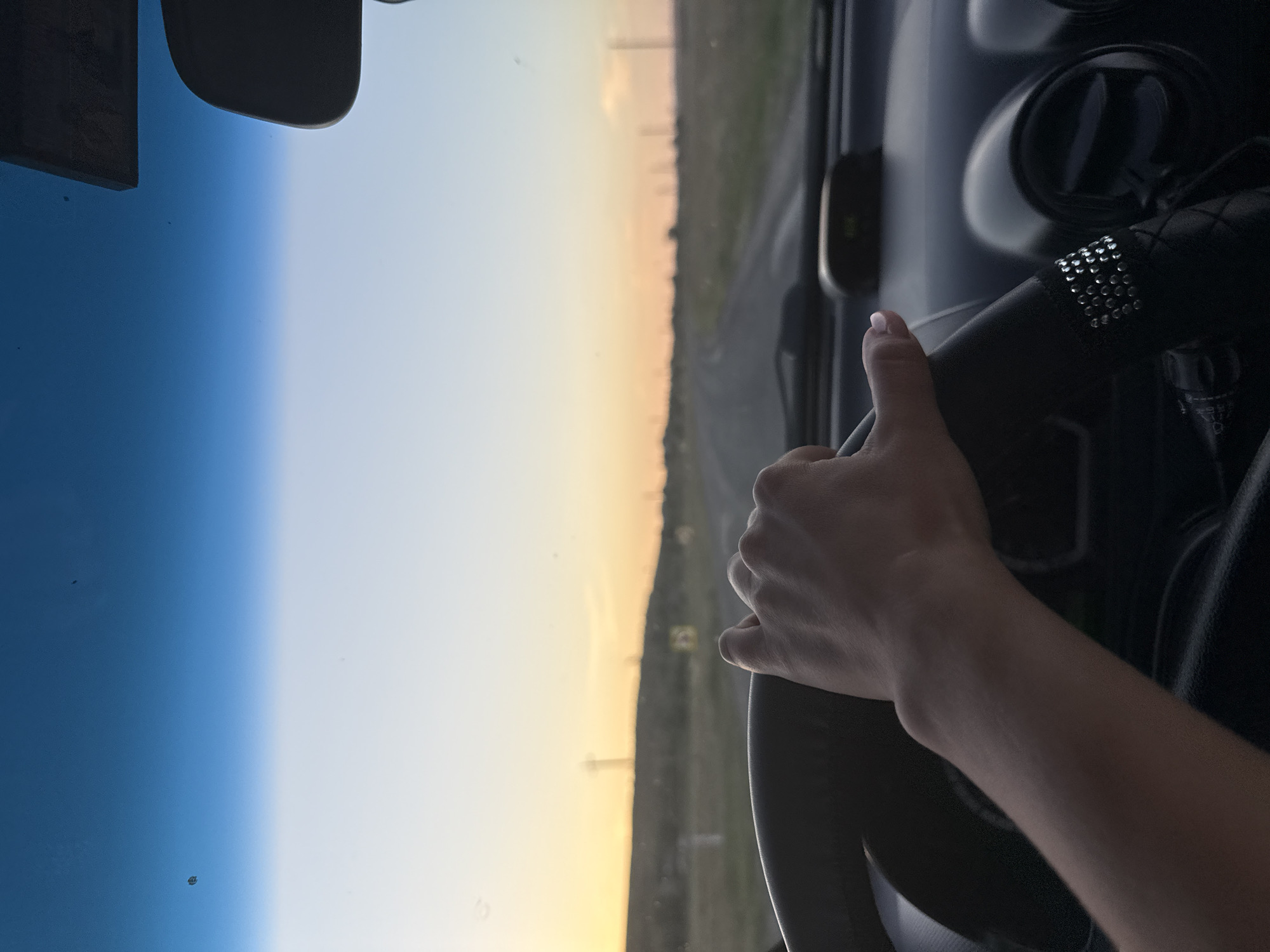 The image shows a first-person perspective of someone driving a car at dusk on a rural road. The sky is a gradient of deep blue at the top fading to orange and yellow near the horizon. The road ahead is visible, lined with sparse vegetation and utility poles. The driver's hand is resting on the steering wheel, which is covered in a black material decorated with rhinestones. The interior of the car is visible, including the dashboard and a rearview mirror. There are a few specks on the windshield. The location is identified as Cioc-Maidan, Moldova.