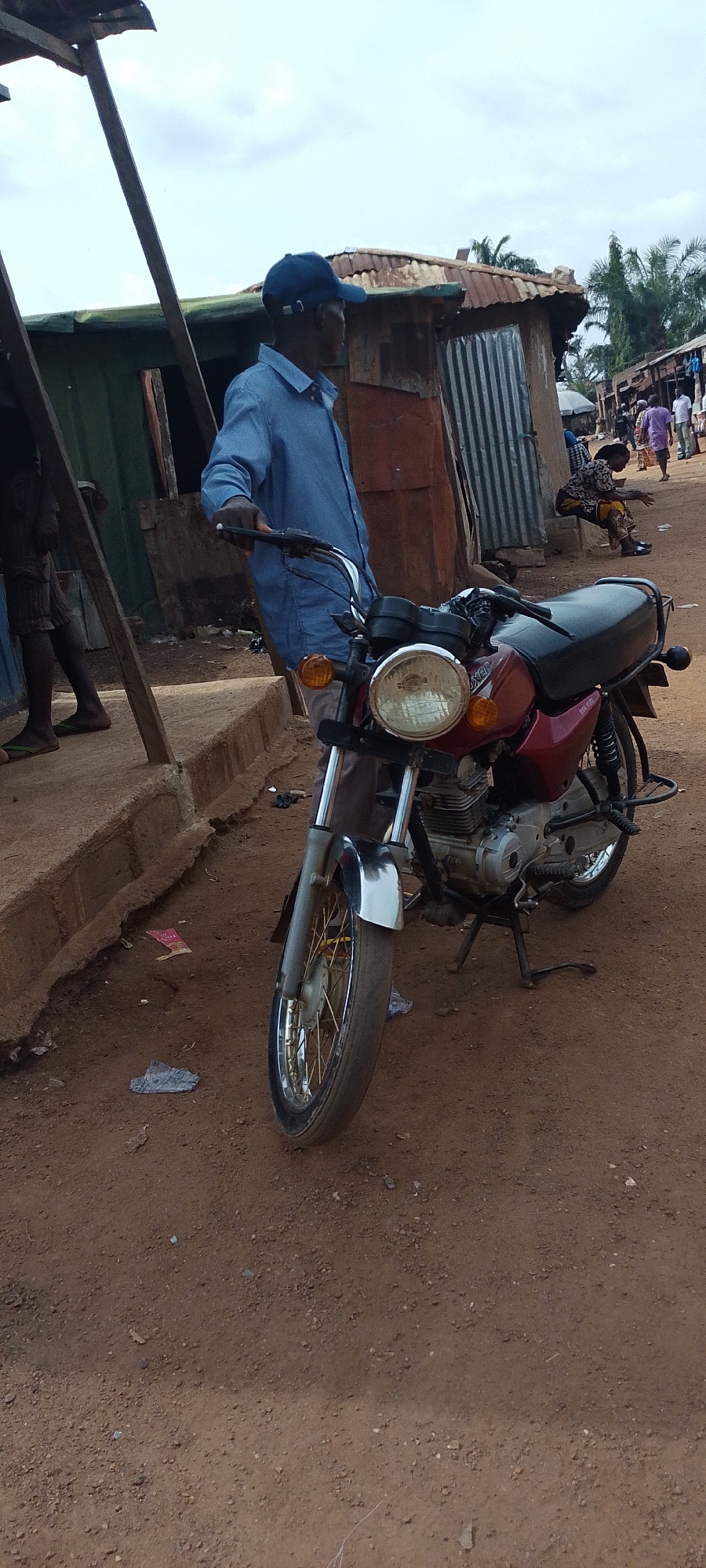 A medium-shot, eye-level, outdoor scene in what appears to be a marketplace or rural area in Gumel, Nigeria, shows a man standing next to a maroon motorcycle. The man, wearing a blue long-sleeved collared shirt and a blue baseball cap, is looking to his right, his left hand on the motorcycle's handlebars. The motorcycle is parked on its kickstand on a dirt ground. In the background, several structures with corrugated metal roofs are visible, suggesting informal housing or market stalls. To the far right, a group of people are walking and standing, with some buildings in the distance. The ground is covered in reddish-brown dirt with scattered debris. The sky is overcast.