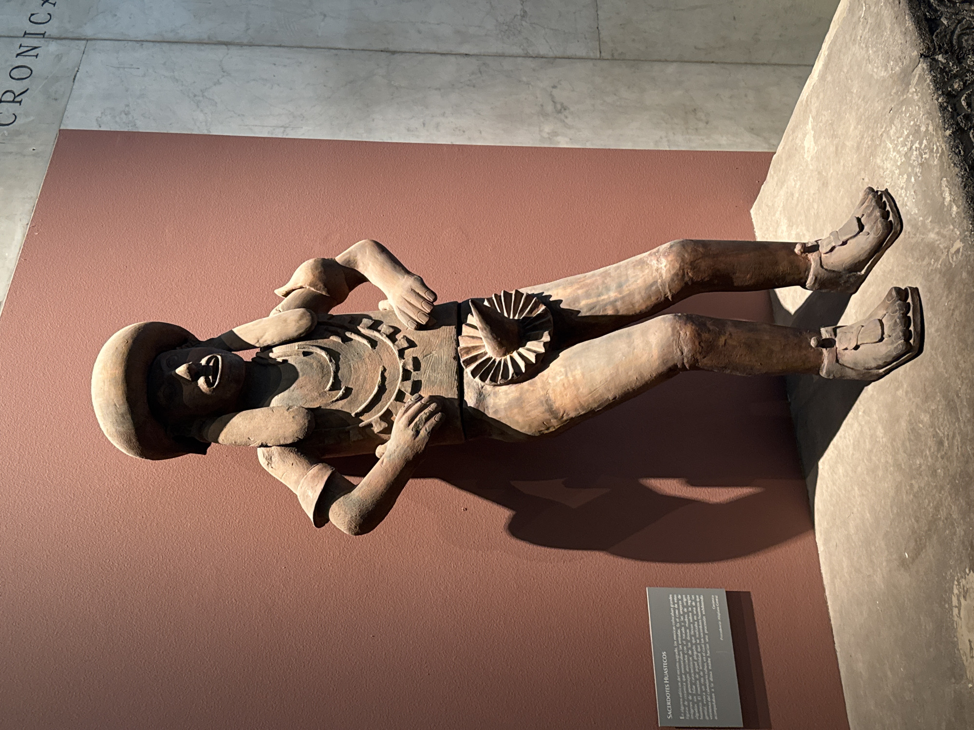 A horizontal, medium shot depicts a terracotta statue of a human figure laying on a reddish-brown surface. The statue is lying on its back, with its legs slightly spread and its arms bent at the elbows. The figure is wearing a hat and sandals. The statue has detailed facial features and clothing. The surface on which the statue is laying is a reddish-brown color. There is a sign in the lower center of the image written in Spanish. The image was taken at an exhibit at the National Museum of Anthropology in Mexico City, Mexico.