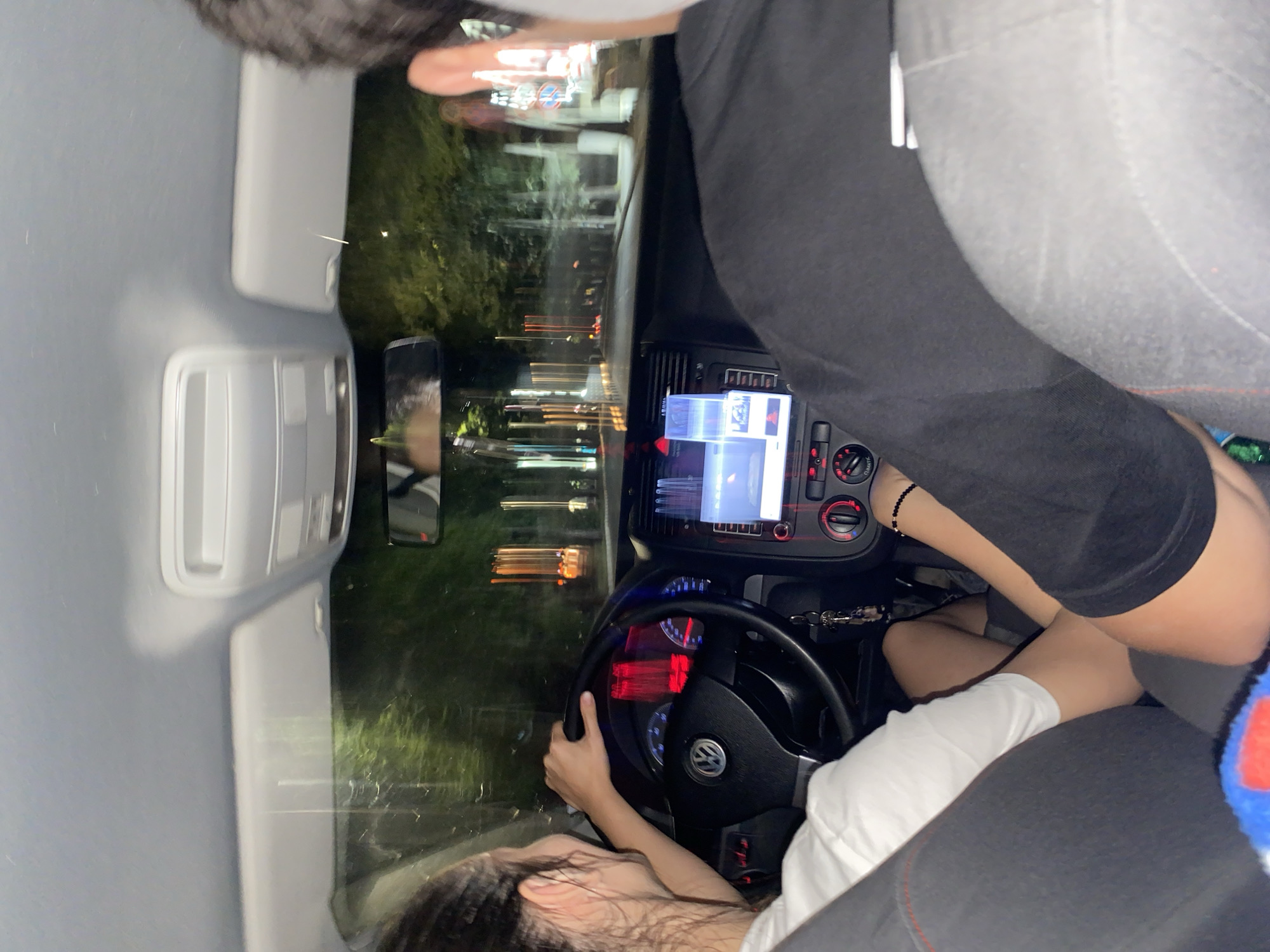 The image shows the interior of a car at night. A woman is driving, her left hand on the steering wheel. She is wearing a white shirt. A passenger is seated to her right, facing forward. They are wearing a black t-shirt. The dashboard shows the car's media system and climate controls. Through the windshield, streetlights and trees are visible, indicating an outdoor urban environment.