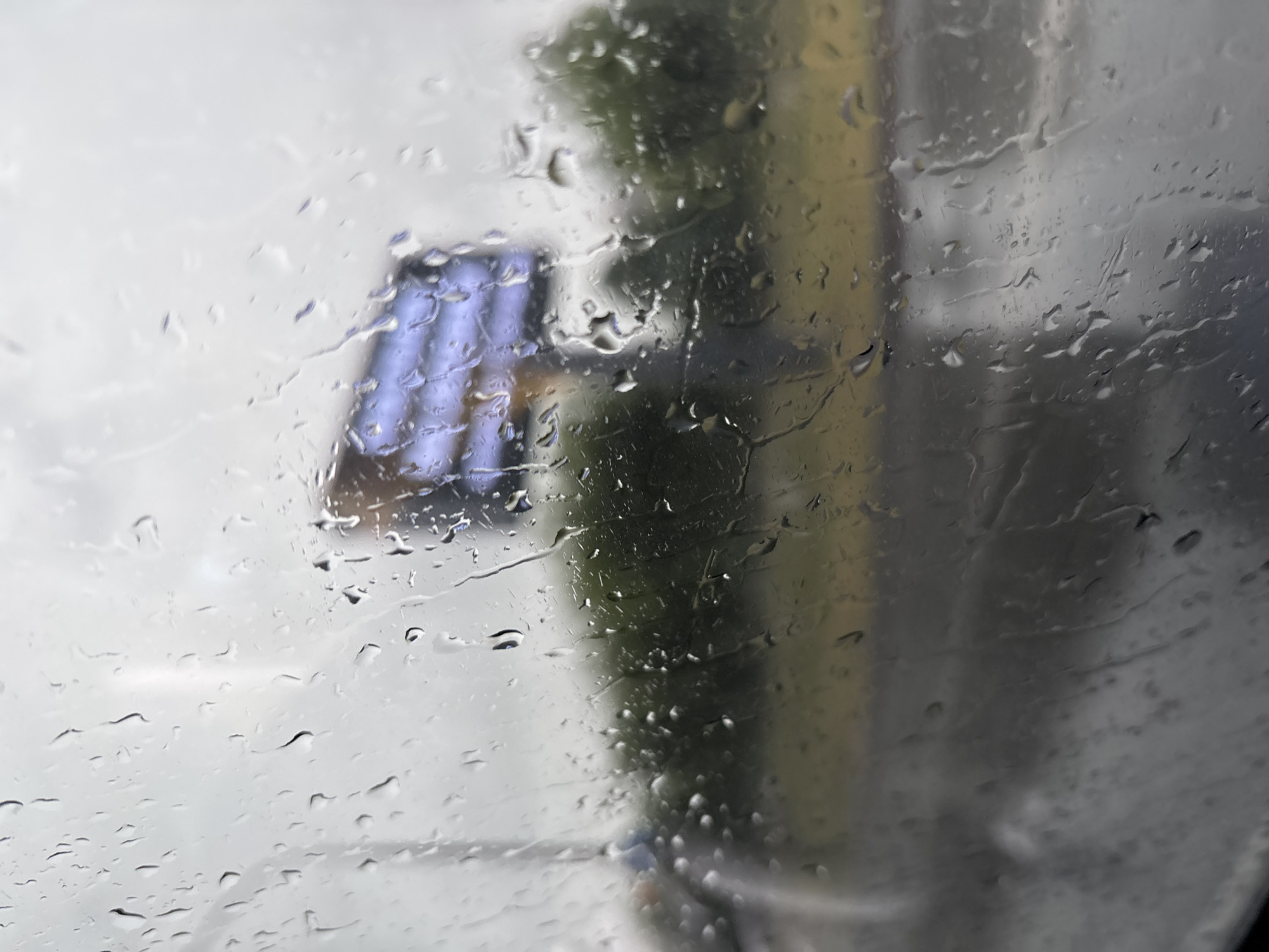 The image captures a view through a rain-streaked window, likely from a vehicle, looking out at a dimly lit, overcast day. In the background, a large, out-of-focus billboard with horizontal blue and white elements is visible. A line of green trees forms a dark band below the billboard, and a blurred, wet road surface is present in the foreground. The presence of rain on the window suggests inclement weather. The location, Naberezhnye Chelny, Russia, does not provide specific visual cues in the image itself, as the scene is generic to many urban or suburban environments during rainy conditions. There are no discernible people, specific objects, or actions occurring that would pinpoint the location further.