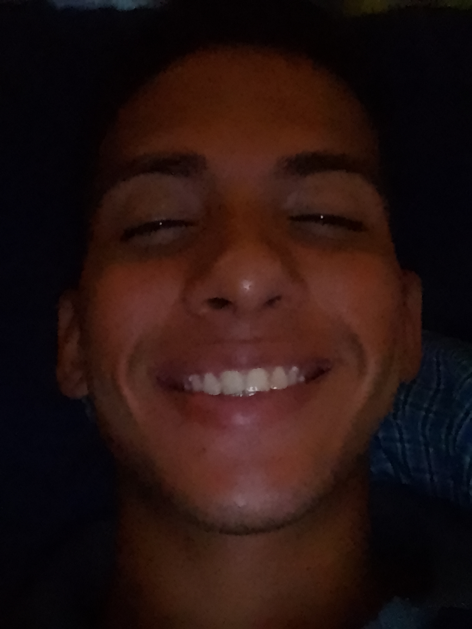 A close-up portrait of a man smiling with his eyes partially closed. He has dark skin, short dark hair, and visible teeth. The background is dark and indistinct, with a blue plaid pattern visible to the lower right of his face. The location, Pampatar, Venezuela, is not discernible from the image. There is no evidence indicating the presence of any objects or specific interactions. The environment is undifferentiated except for the plaid cloth.
