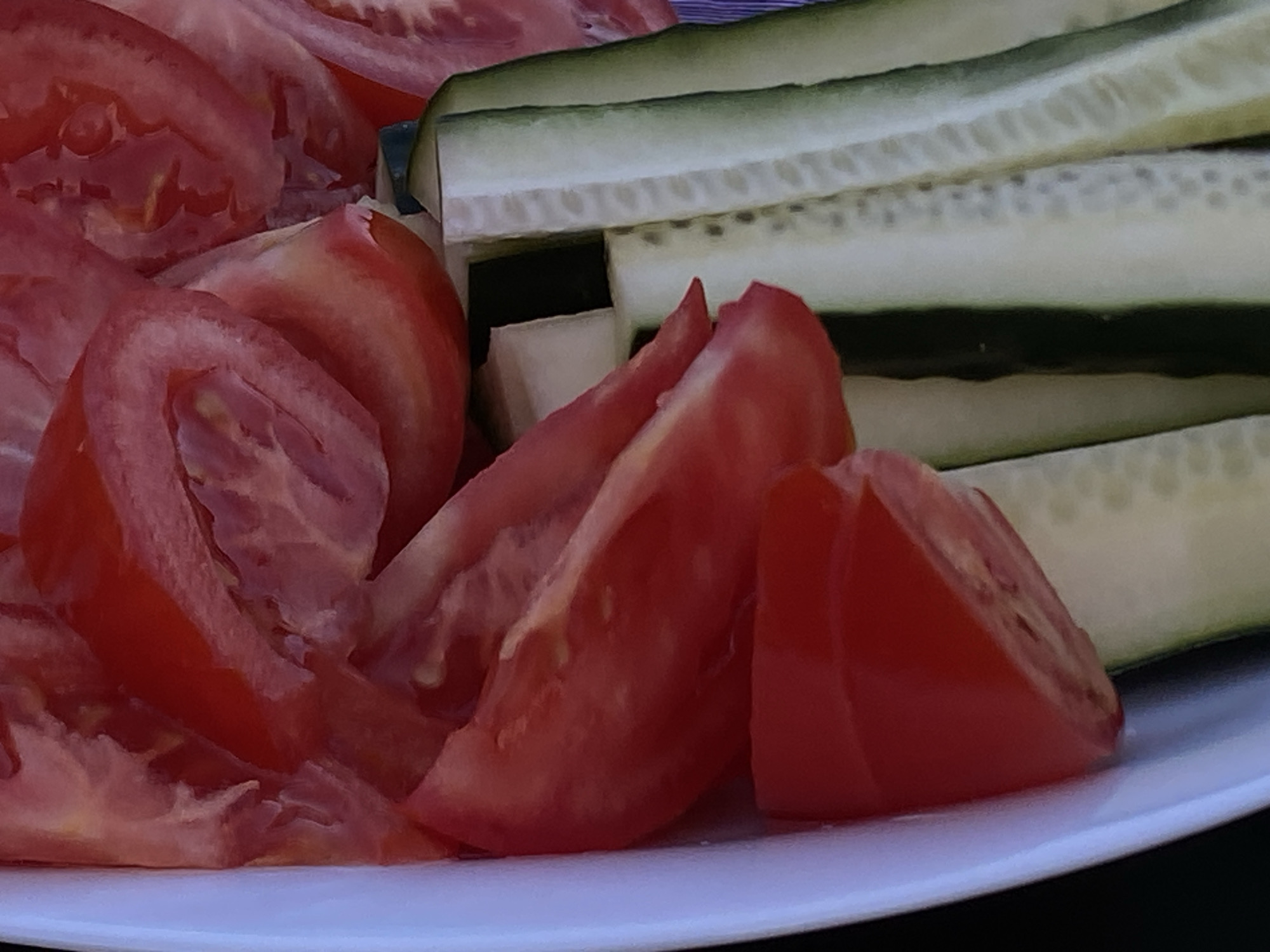 A close-up, overhead shot captures sliced tomatoes and cucumbers on a white plate, with a dark background. The tomatoes are cut into wedges and arranged on the left side of the plate, showcasing their vibrant red flesh and seeds. The cucumbers are sliced thinly and stacked in layers on the right, revealing their pale green skin and crisp white interiors. The lighting is soft and natural, highlighting the fresh textures of the vegetables. The scene is static and appears to be indoors or in a shaded outdoor area, given the lack of harsh shadows. The image has a shallow depth of field, with the foreground elements sharp and the background slightly blurred. There is no indication of the location being Arandjelovac Municipality, Serbia, based on the visual information provided.