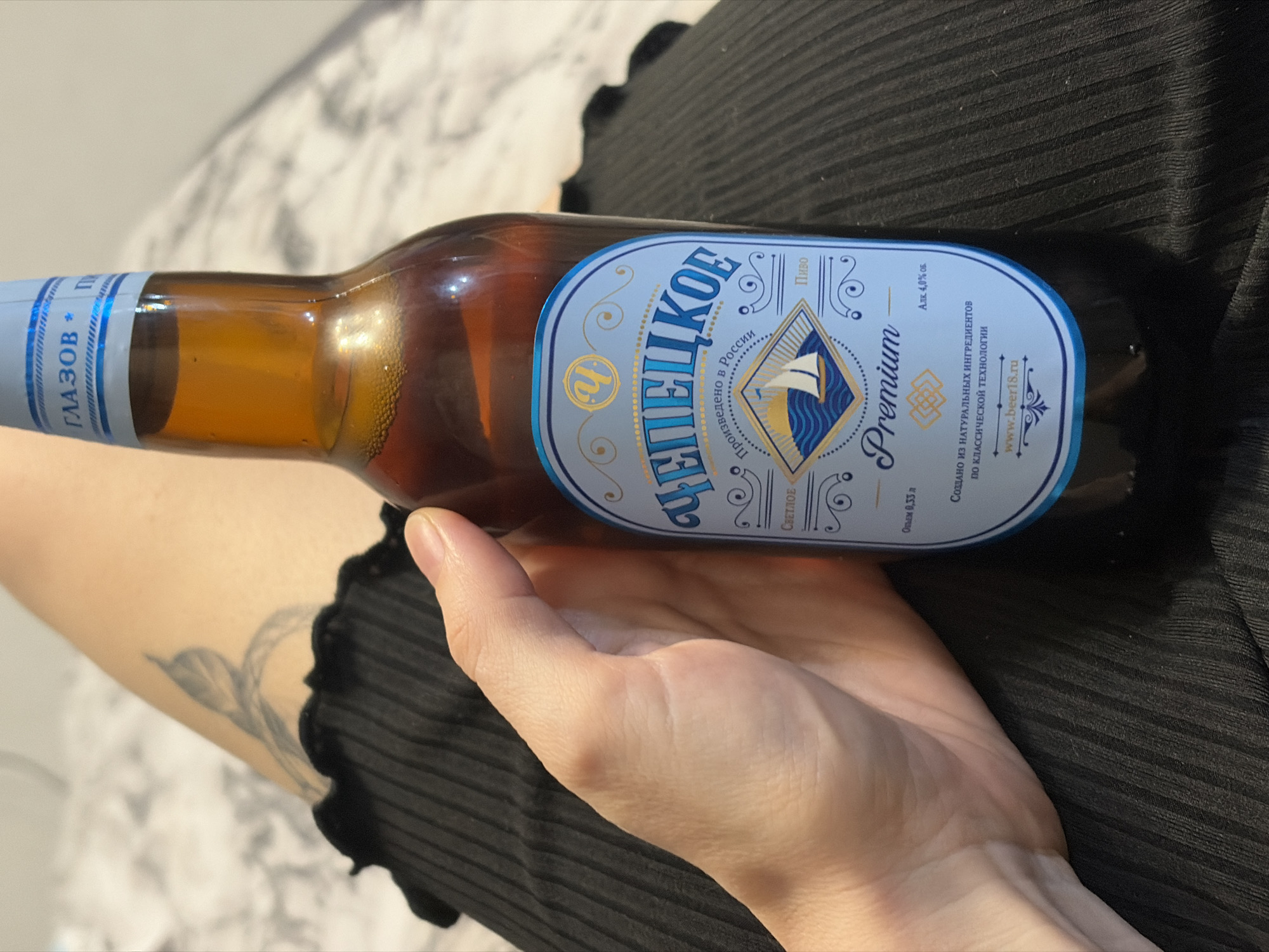 A hand is holding a brown glass bottle of "Chepetskoe" Premium beer. The bottle's label states that it is produced in Russia, is a "light" beer, contains 4.0% alcohol by volume, and is created from natural ingredients using classic technology. The label also includes a website, "www.beer18.ru". "Glazov" is written on the neck label. The bottle rests against the person's leg, which is covered in black fabric with a ribbed or textured pattern. Part of the person's arm is visible to the left, showing a tattoo of a plant or abstract shape. The background is blurry, showing a pattern that could be a bedspread or similar textile.