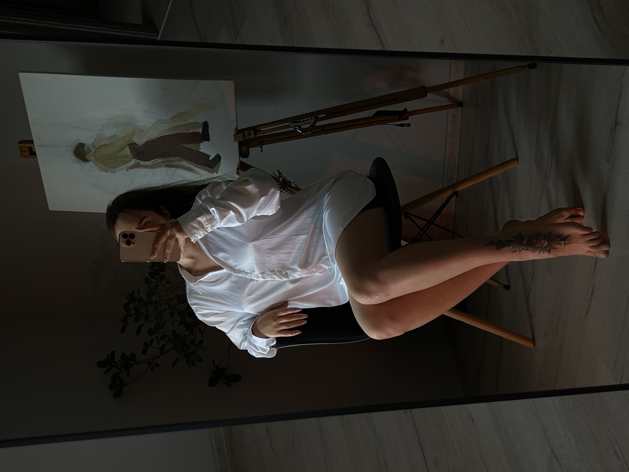 A young woman is seated on a black chair, holding a smartphone to take a mirror selfie. She is wearing a loose, white button-down shirt, her legs are crossed, and she has a floral tattoo on her left ankle. In the background, an easel holds a partially completed painting of two figures. The room appears to be an artist's studio, with wooden flooring and a large mirror reflecting the scene. The lighting is dim, with a strong spotlight coming from the right, creating dramatic shadows. The image was taken in Ternopil, Ukraine.
