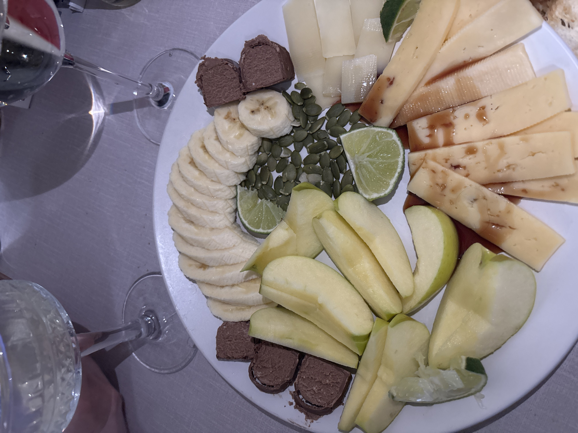 The image displays a top-down view of a white round plate filled with various snacks, accompanied by two stemmed glasses. The setting appears to be indoors, likely a dining table, under artificial lighting, suggesting an evening occasion.
On the plate, an assortment of foods is arranged. Towards the upper left, there are several irregular chocolate candies or truffles. Below them, a fanned arrangement of peeled banana slices forms a semi-circle. Adjacent to the bananas and extending into the center are numerous green pumpkin seeds. Several bright green lime wedges are interspersed across the plate.
On the right side of the plate, various cheeses are presented. Some are light-yellow block slices, while others are longer, thinner slices drizzled with a dark, reddish-brown sauce or syrup. Below the pumpkin seeds and limes, a collection of light-green apple slices, some peeled, fill the lower right portion of the plate.
Two stemmed glasses are visible outside the plate's perimeter. The glass in the upper left contains a dark red liquid, indicative of red wine, and shows reflections. Another glass is partially visible at the bottom, appearing to hold a lighter liquid. The surface beneath the plate and glasses is a light-colored, textured material, possibly a tablecloth. The overall presentation suggests a thoughtful snack or dessert platter for sharing, indicative of a relaxed social gathering in Perm, Russia. No visible text is present.
