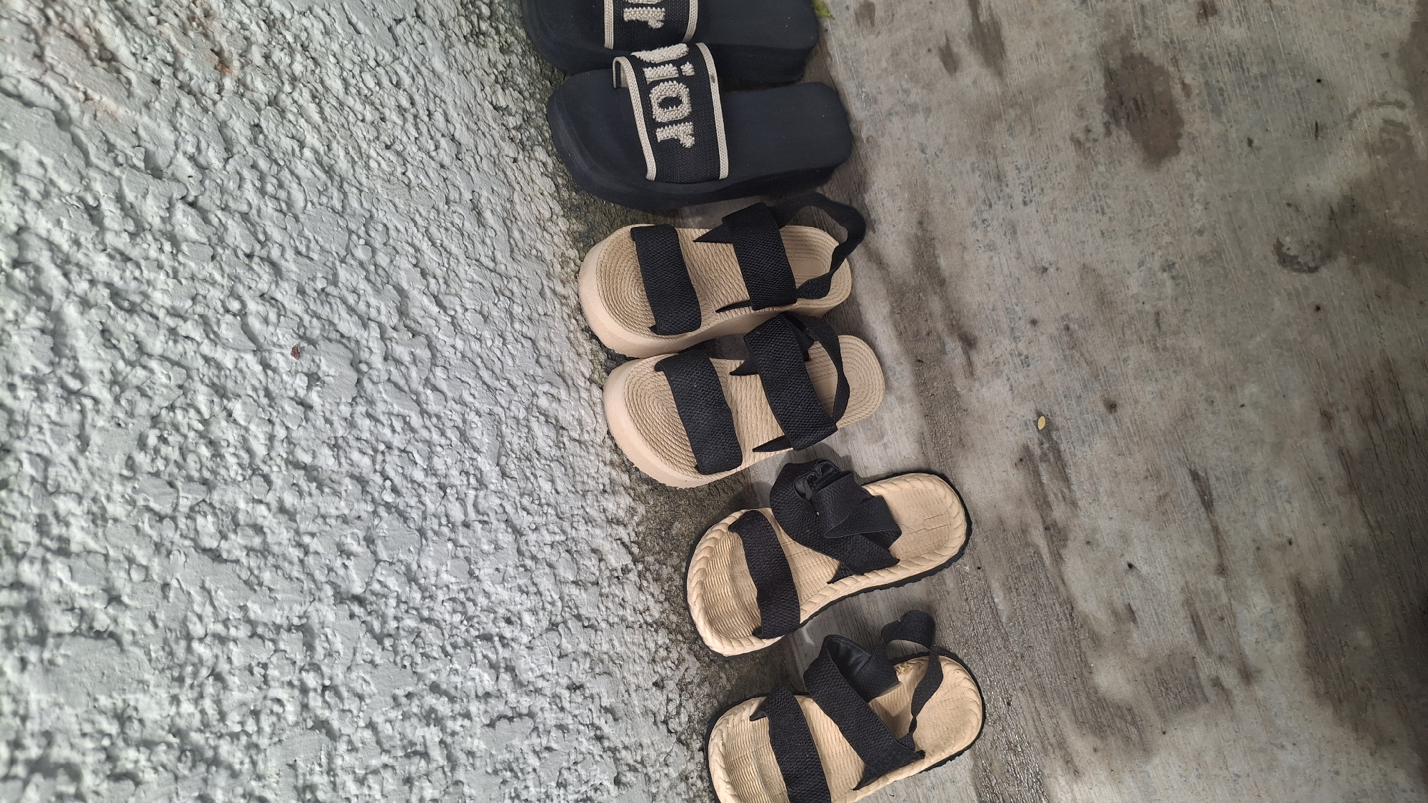 The image displays a close-up, eye-level view of five pairs of sandals lined up on a weathered wooden surface. The sandals are positioned against a rough, textured, pale grey wall.
From left to right, there are two beige sandals with black straps. The first sandal on the far left has its sole visible and appears to be a men's or women's walking sandal. Next to it is another similar beige sandal, slightly angled.
Following these are two more beige sandals with black straps, appearing to be a matching pair, placed side-by-side. These sandals have a thicker sole than the first two.
On the far right, there are two black slide-style sandals. The first of these has a white strap with "DIOR" written in black lettering. The second black sandal is mostly obscured by the first.
The wooden surface shows signs of wear, with some water stains and lighter patches. The wall behind is textured with an uneven, pebbly finish. There are no people or animals visible in the scene. The lighting suggests it is daytime, possibly overcast, with diffused light creating soft shadows. The setting appears to be an outdoor or semi-outdoor area, common in residential or commercial spaces in Jalingo, Nigeria, where footwear is often removed before entering or left outside.