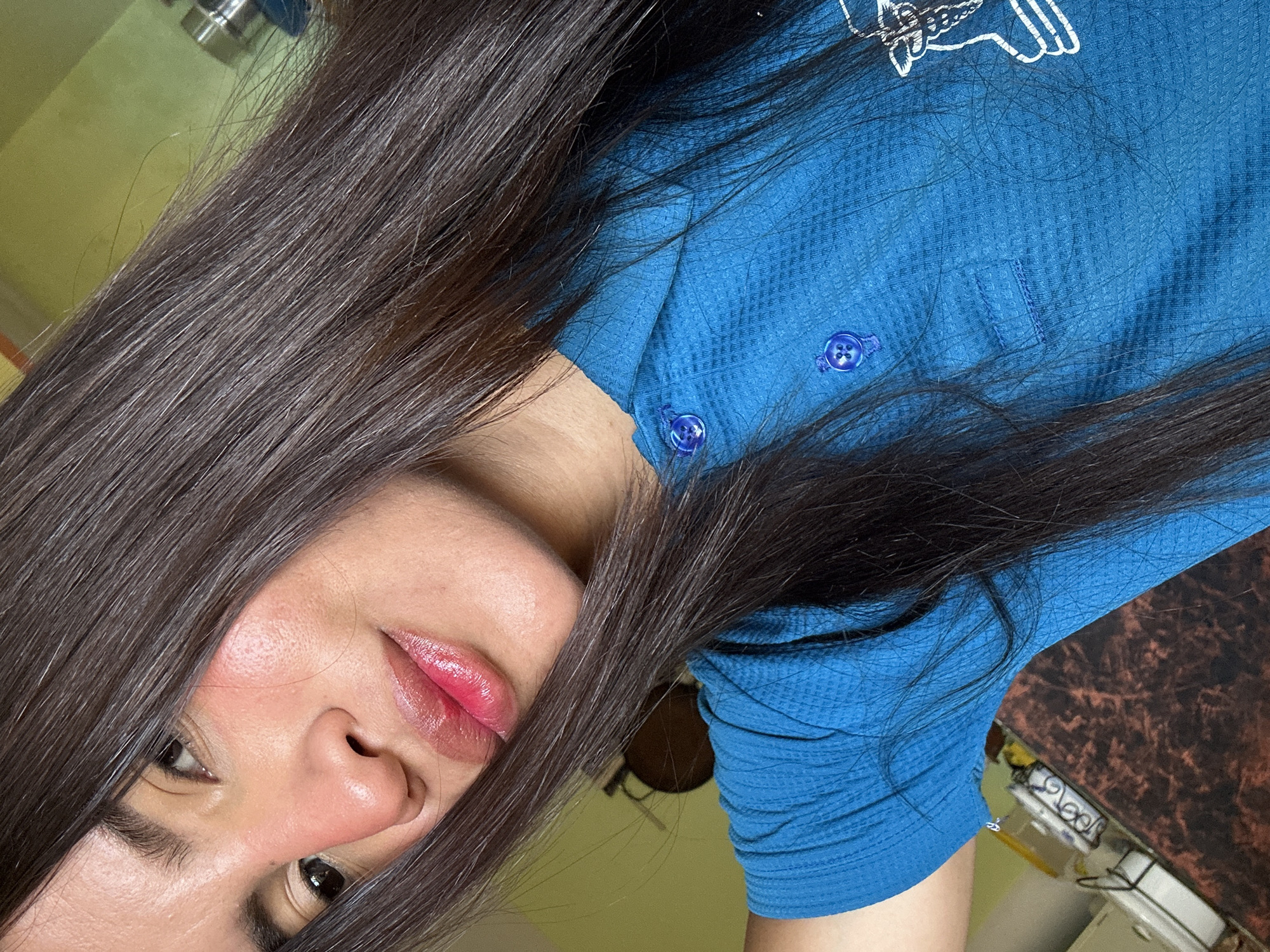 The media file is a close-up, waist-up shot of a young woman with long, dark hair that partially obscures her face. She is wearing a blue textured shirt with two visible navy blue buttons. Her lips are painted a bright pink and appear slightly glossy. Her skin is fair with a hint of blush on her cheeks, and her eyes are dark. The image is taken indoors, with a light yellow wall and a dark brown patterned surface visible in the background. The overall lighting suggests it is daytime. There is no visible text in the media file. The composition and lighting suggest a casual, personal photo.