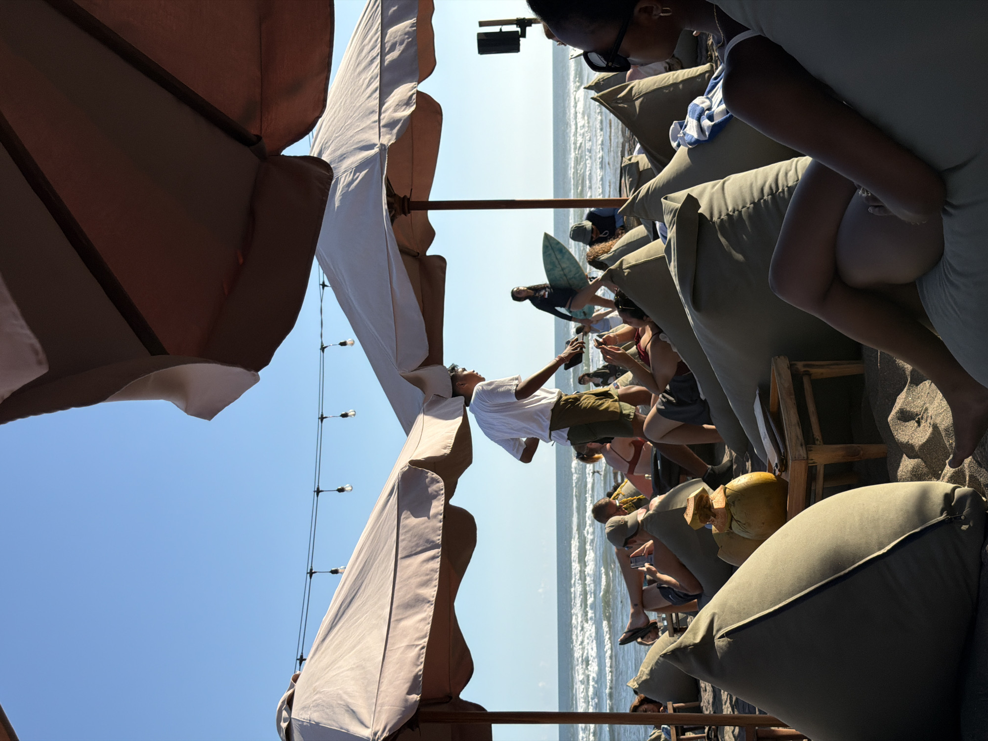 This media file depicts a group of people relaxing on a beach, likely at a resort or beach club, with a view of the ocean. The scene is set under large, light-colored umbrellas, providing shade. Several individuals are visible, seated or lounging on large, cushioned beanbag-like chairs. Some people are looking at their phones, while others are engaged in conversation or simply enjoying the ambiance.
A young man in a white t-shirt and olive green shorts is prominently featured, seemingly posing or balancing with his arms outstretched. Another individual is holding a surfboard. The ocean is visible in the background, with gentle waves and the horizon. String lights are also visible, suggesting that this could be an area used during the evening as well. The sand and the presence of umbrellas and beach furniture strongly indicate a beachside location.
The time of day appears to be daytime, with bright sunlight casting shadows. The weather seems pleasant and sunny, suitable for a beach outing. While no text is explicitly visible on clothing or objects, the overall atmosphere suggests a relaxed and possibly vacation-like setting. The location context of North Kuta, Indonesia, further supports the interpretation of a tropical beach scene.