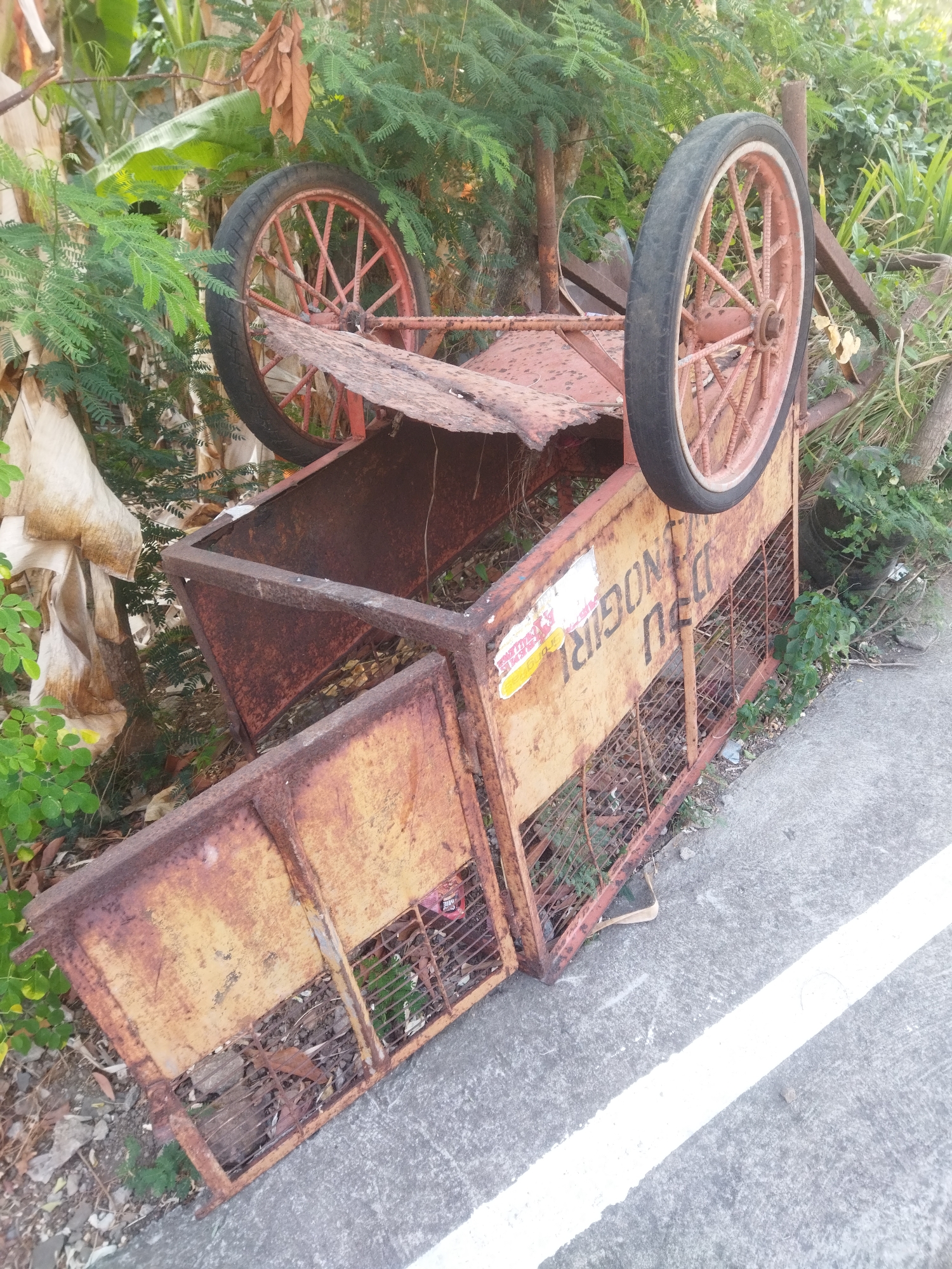 The image shows an overturned, rusty, wheelbarrow-like cart with two spoked wheels on a roadside in Wonogiri, Indonesia. The cart appears to be made of metal, with a yellow and rust-colored body. There are some green plants and trees in the background, and a concrete road with a white line runs diagonally across the foreground. The cart is positioned near some foliage, with its wheels facing upwards. There are no people visible in the image. The scene suggests the cart may have been involved in an accident or has been abandoned.