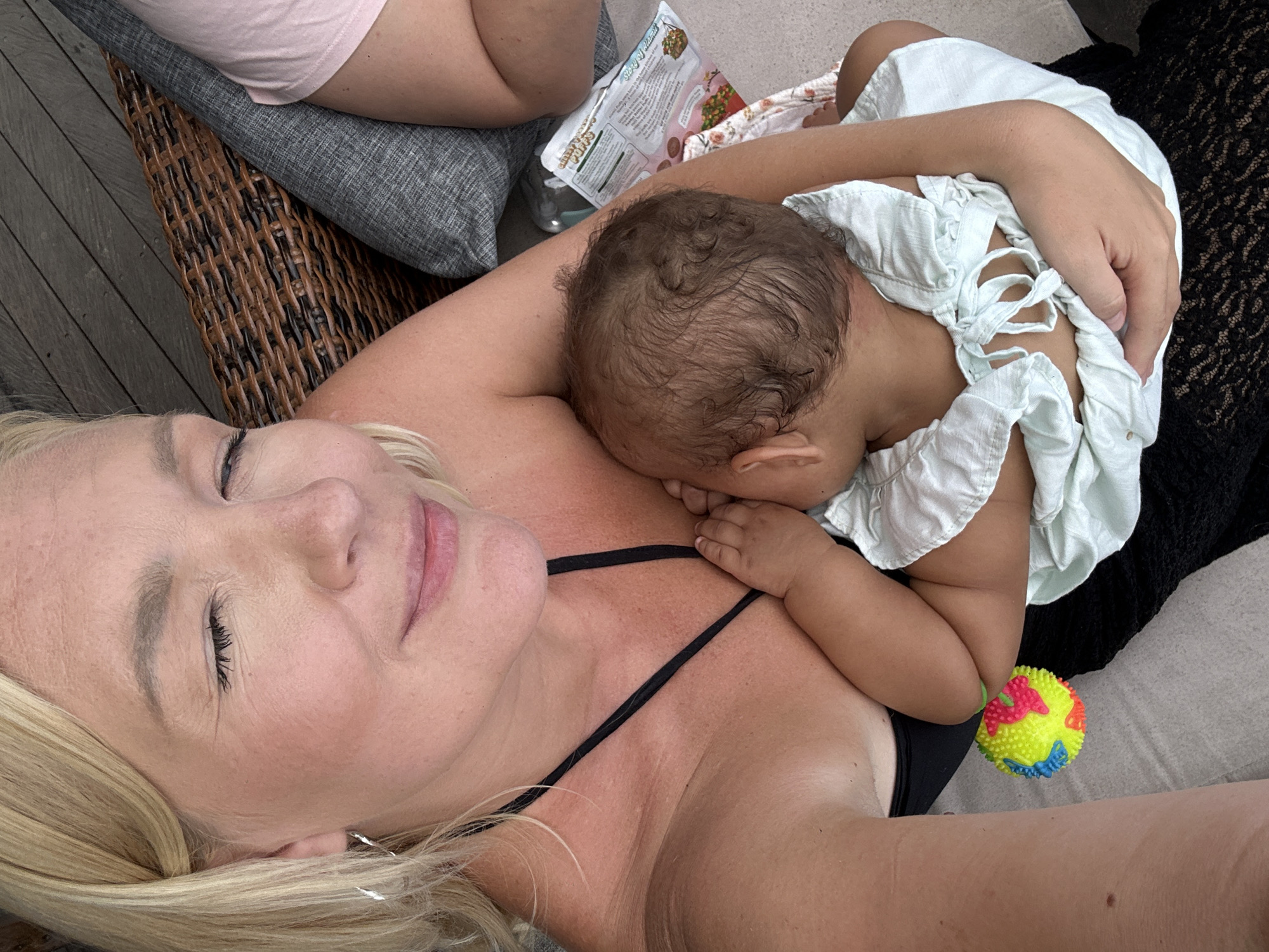 A medium-shot, eye-level photograph shows a fair-skinned woman lying on her back with an infant asleep on her chest. The woman has blonde hair and closed eyes, and is wearing a black-strapped top. The infant has dark curly hair and is wearing a light-colored top with ties at the back. A colorful ball is positioned near the infant’s legs. Part of another person’s legs and a grey pillow are visible in the upper-left corner of the image. Also visible in the background is a wicker object, a clear bottle, and a package with text on it. The location is identified as North Kuta, Indonesia.