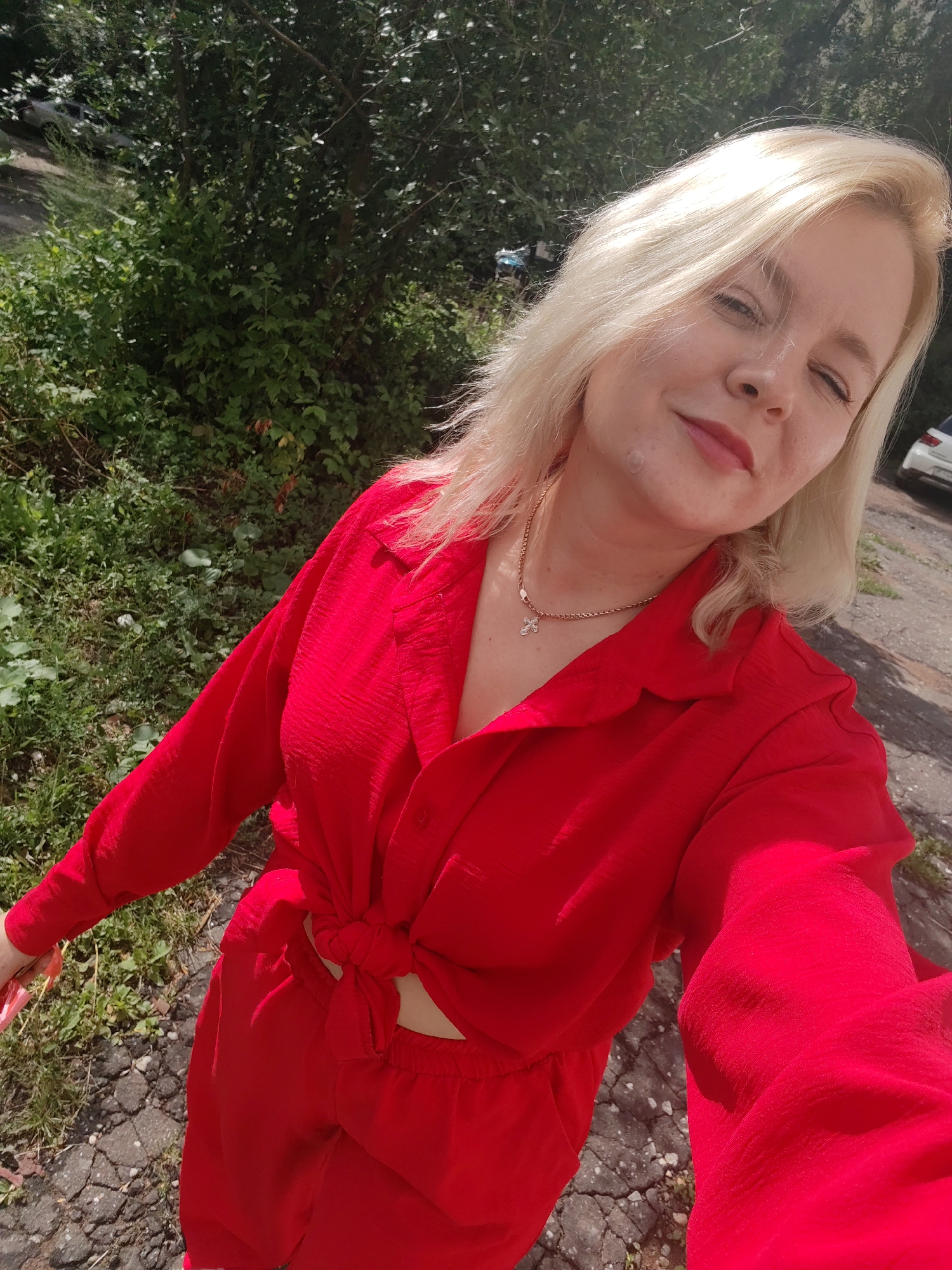 A woman with blonde hair is taking a selfie outdoors. She is wearing a red shirt tied at the waist and red pants. She has a cross necklace on. In the background, there are trees and a car. The ground is paved with cracked asphalt and there is some grass. The overall impression is that of a sunny day. The image appears to have been taken in Izhevsk, Russia, based on the provided context.