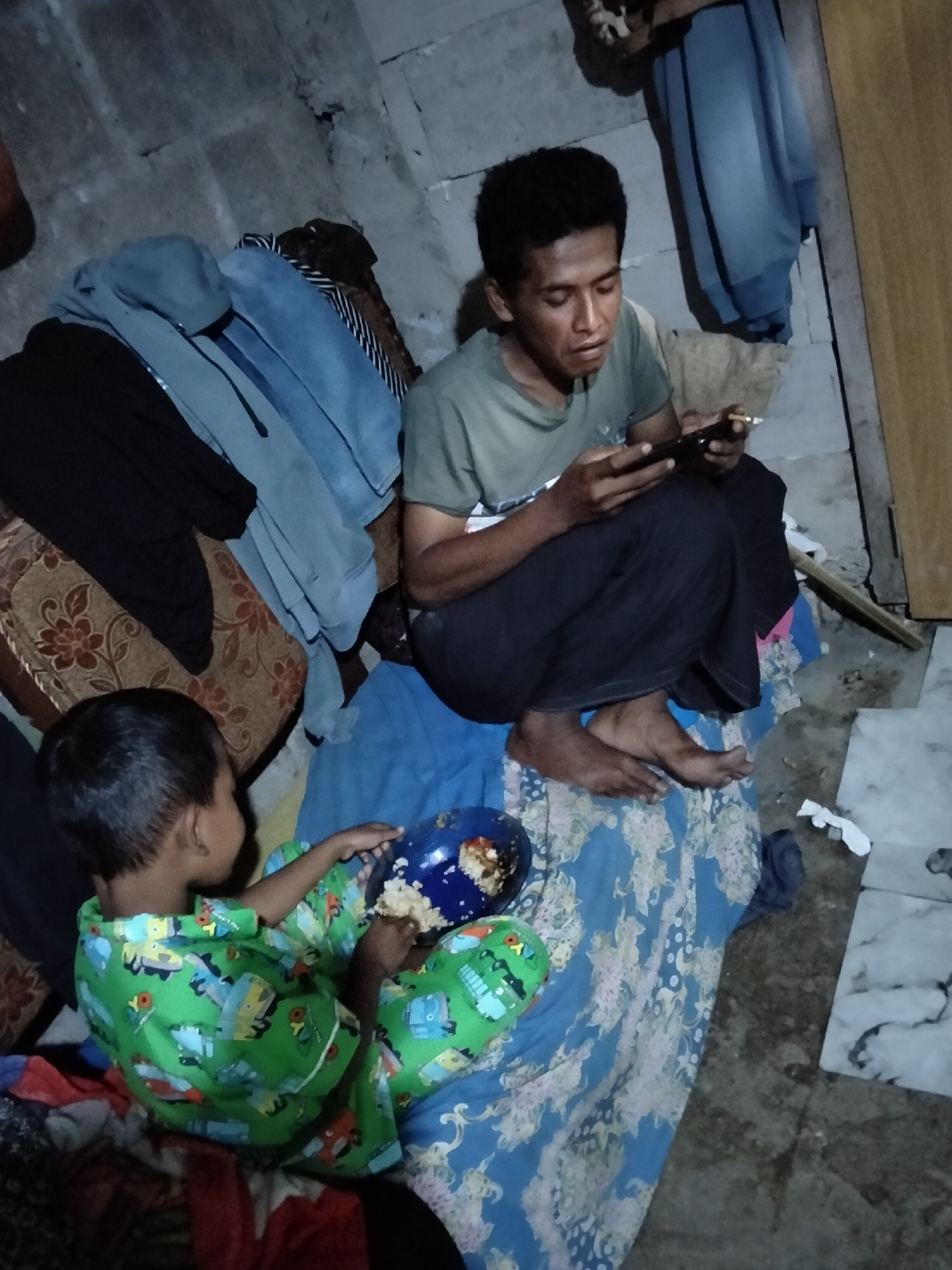 In Puspo, Indonesia, a man and a young child are on the floor in what appears to be a living space. The man, wearing a green t-shirt and dark pants, is seated and looking at a mobile phone. The child, dressed in a green pajama set with cartoon vehicle prints, is seated on a blue and floral patterned blanket and is holding a blue bowl with food. Scattered around them are various clothing items, including a black jacket, blue sweaters, and patterned fabrics, suggesting the area is used for storage or is in a state of disarray. The floor is concrete, and the walls are rough and textured. A blue garment is draped over a structure in the background. The lighting is dim, indicating an indoor setting possibly at night or in a poorly lit area.