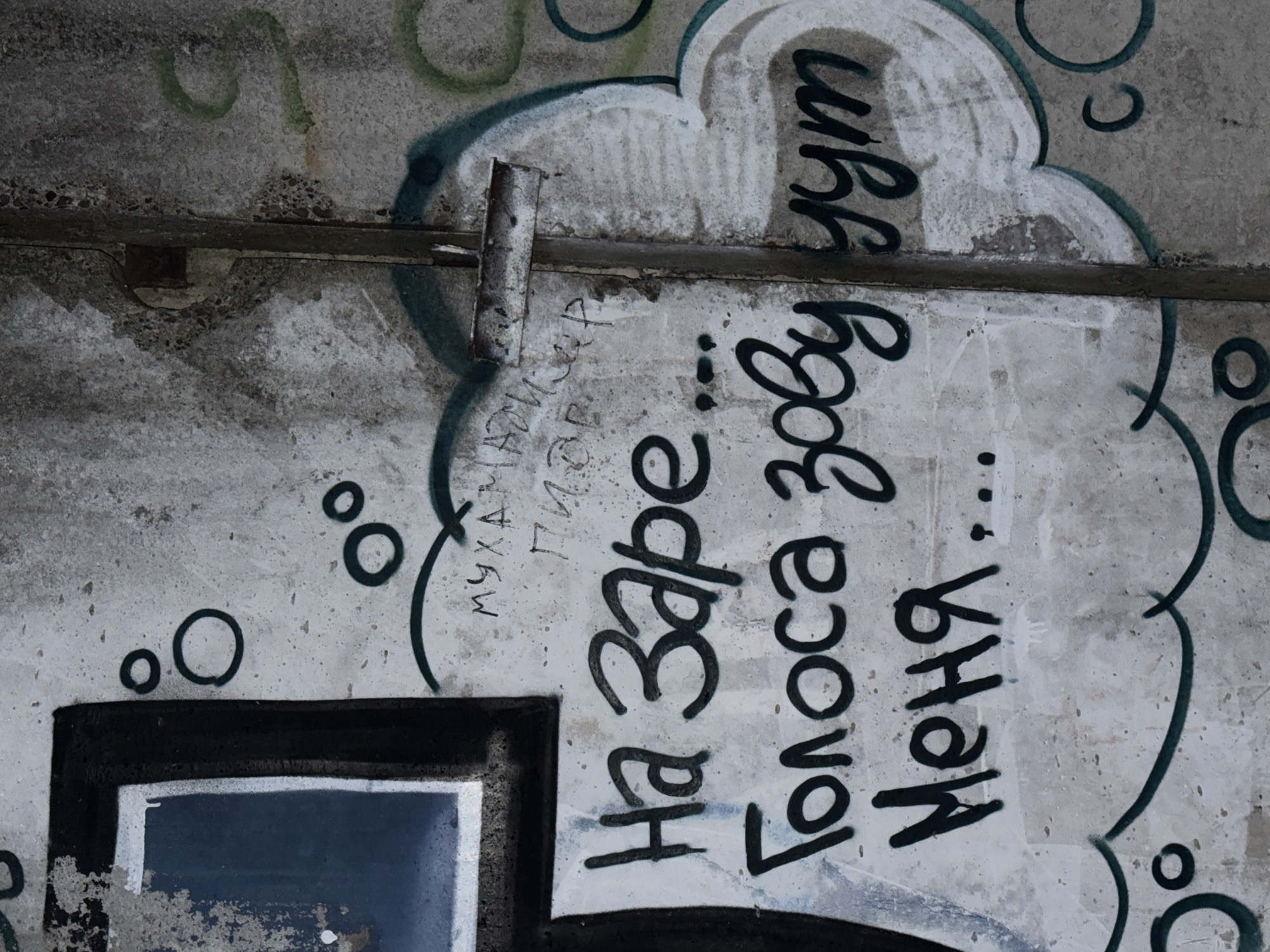 A close-up shot reveals a concrete wall covered in graffiti. The wall's surface is mottled with patches of discoloration and wear, suggesting age and exposure to the elements. The graffiti appears in dark paint and features a mix of Cyrillic letters and symbols, possibly indicating tags or street art. The script is in Russian: "На Заре... Голоса зову меня..." which translates to "At Zare... Voices are calling me..." The shot also includes a metallic, cylindrical object affixed to the wall, further adding to the urban decay aesthetic. A partially visible dark square, potentially a window or some other architectural feature, is seen at the lower left corner. The location is the city of Chaikovsky, Russia.