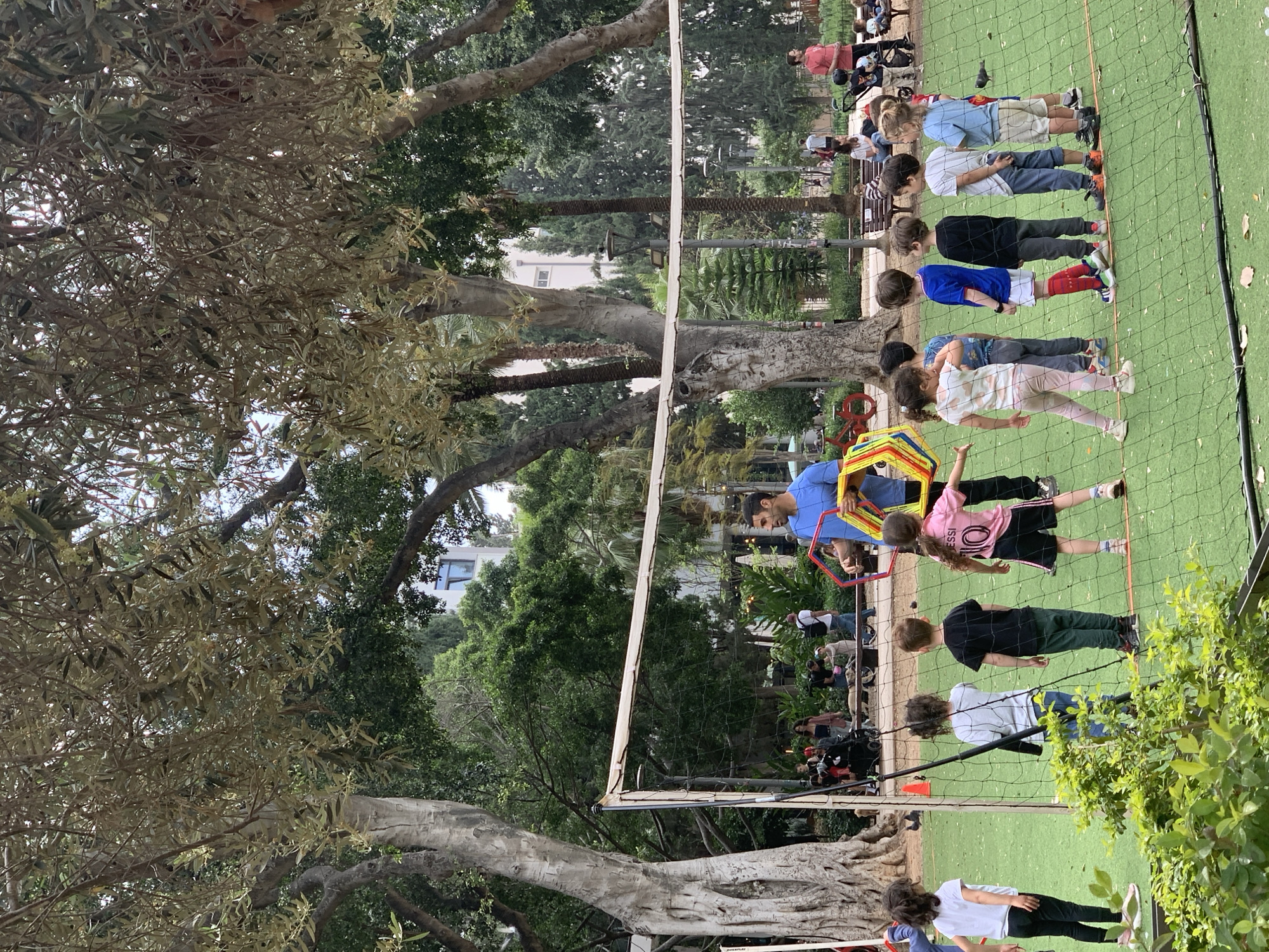 A group of children are participating in an activity at a playground or park with a net structure, likely near a park in Tel Aviv, Israel, suggested by the lush greenery and type of trees present. The scene appears to be outdoors on a sunny day. A volleyball net is prominently featured, running across the middle ground of the frame. The ground is covered in artificial turf.
On one side of the net, a row of children stands in line, seemingly engaged in some sort of organized game or exercise. A man, presumably an instructor, is assisting and guiding a child through a hula hoop-like structure. Several other people are visible in the background, suggesting the presence of supervising adults.
The environment consists of a mix of trees, including palm trees and other species common to the Mediterranean region. Some buildings are visible in the background, possibly residential or community structures.