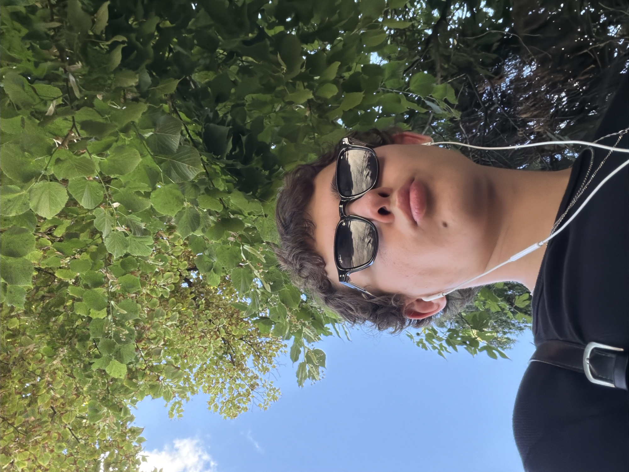 An individual is depicted from a low-angle perspective, captured in a close-up, outdoor setting within Palilula Urban Municipality, Serbia. The person has short, curly dark hair and wears dark-framed sunglasses with highly reflective lenses, which mirror the sky and surrounding foliage. White wired earphones are inserted into both ears, with the wires trailing downwards. The individual's lips are distinctly puckered. A small dark mark is present on the skin below the right side of the lower lip. The person is wearing a black top or t-shirt, and a silver-colored chain necklace is visible around the neck. A black strap with a silver rectangular buckle is visible over the left shoulder, consistent with a bag strap. The background features a vibrant blue sky with scattered white clouds in the upper-left portion of the frame, transitioning to a dense canopy of green tree leaves that dominate the upper and right sections of the image. The leaves are broad, ovate, and well-lit by natural daylight.