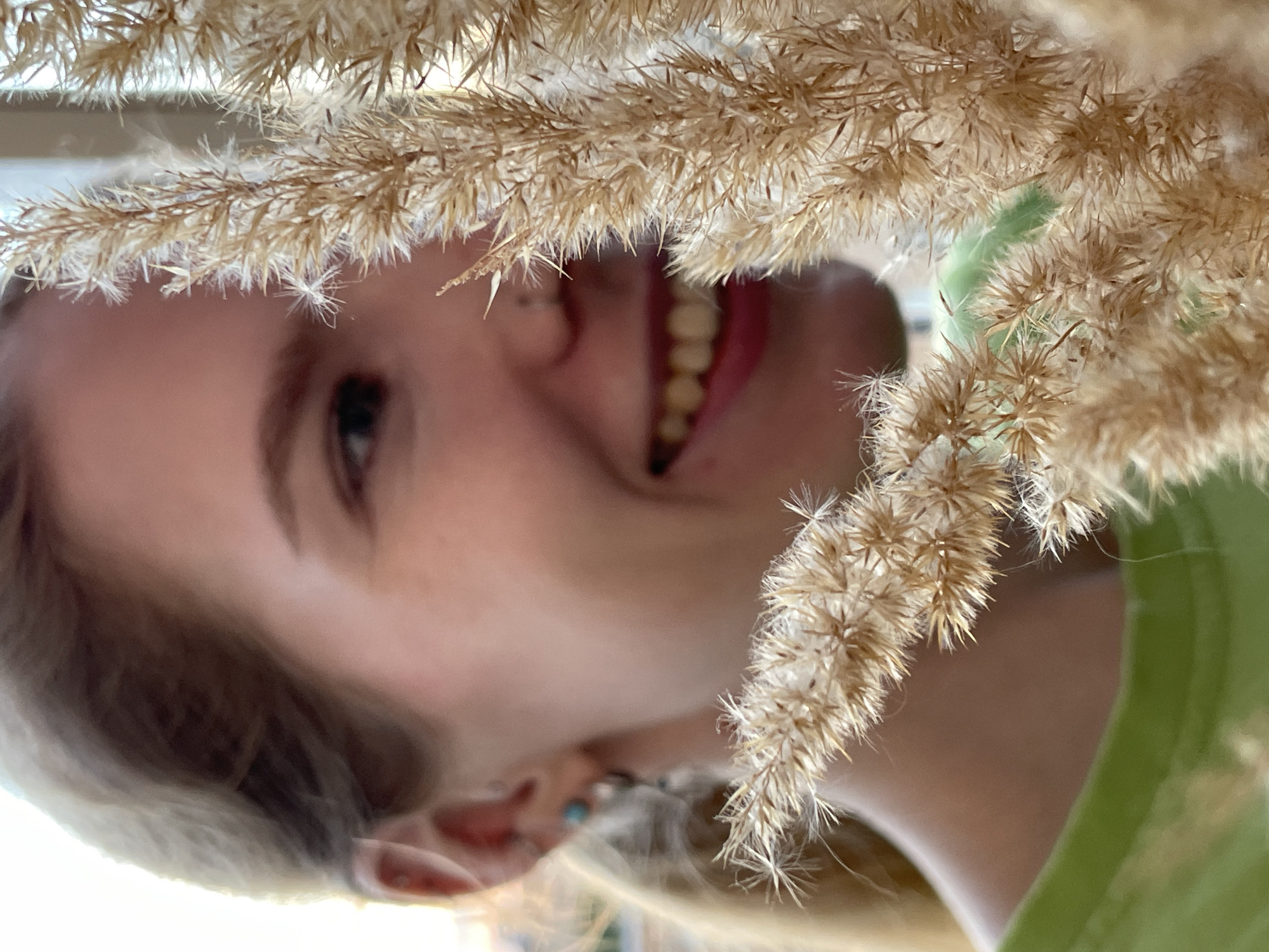 A young fair-skinned woman with blonde hair is partially visible behind a bunch of dried wheat-like grass. She is smiling, revealing her teeth. She has a nose piercing and is wearing earrings. A green shirt is visible. The location is unspecified and there is no evidence to suggest it is in Izhevsk, Russia.