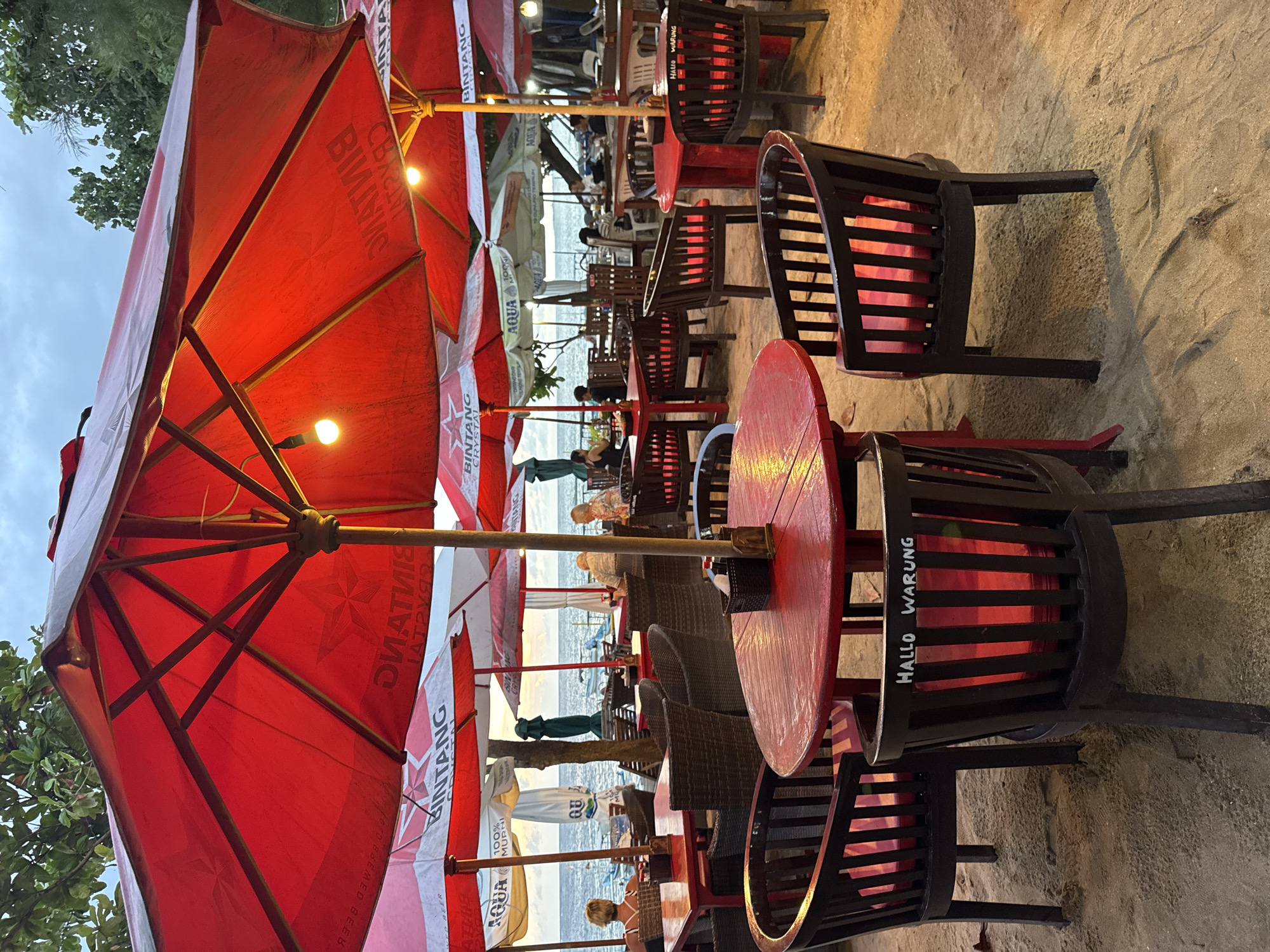The image depicts an outdoor dining area on a beach in Denpasar, Indonesia. Several red and white umbrellas with the logos "BINTANG," "AQUA," and partially obscured text shield the tables from the sun. The tables are round and painted red, surrounded by black wooden chairs with "HALLO WARUNG" painted on them. People are seated at some tables, enjoying the view of the ocean in the background. The ground is sandy. The overall lighting suggests it's either early morning or late afternoon.