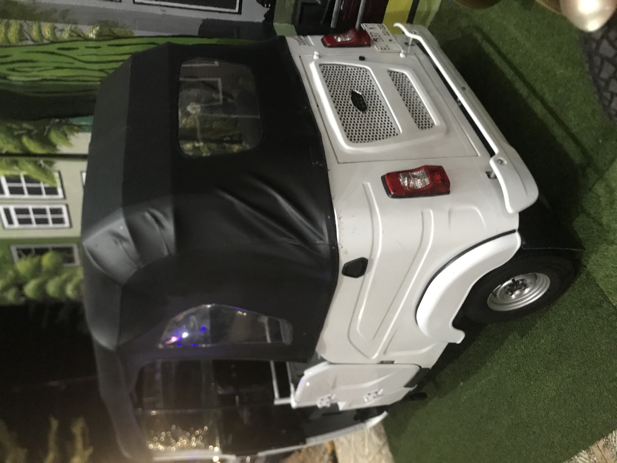 The image shows a three-wheeled vehicle, possibly an auto rickshaw or "tuk-tuk," with a white body and a black canvas top. It is parked on what appears to be artificial turf resembling grass. The vehicle's rear lights and grill are visible. In the background, there is a blurred image of a building façade with windows and greenery. The location is Keudeteupinpunti, Indonesia.