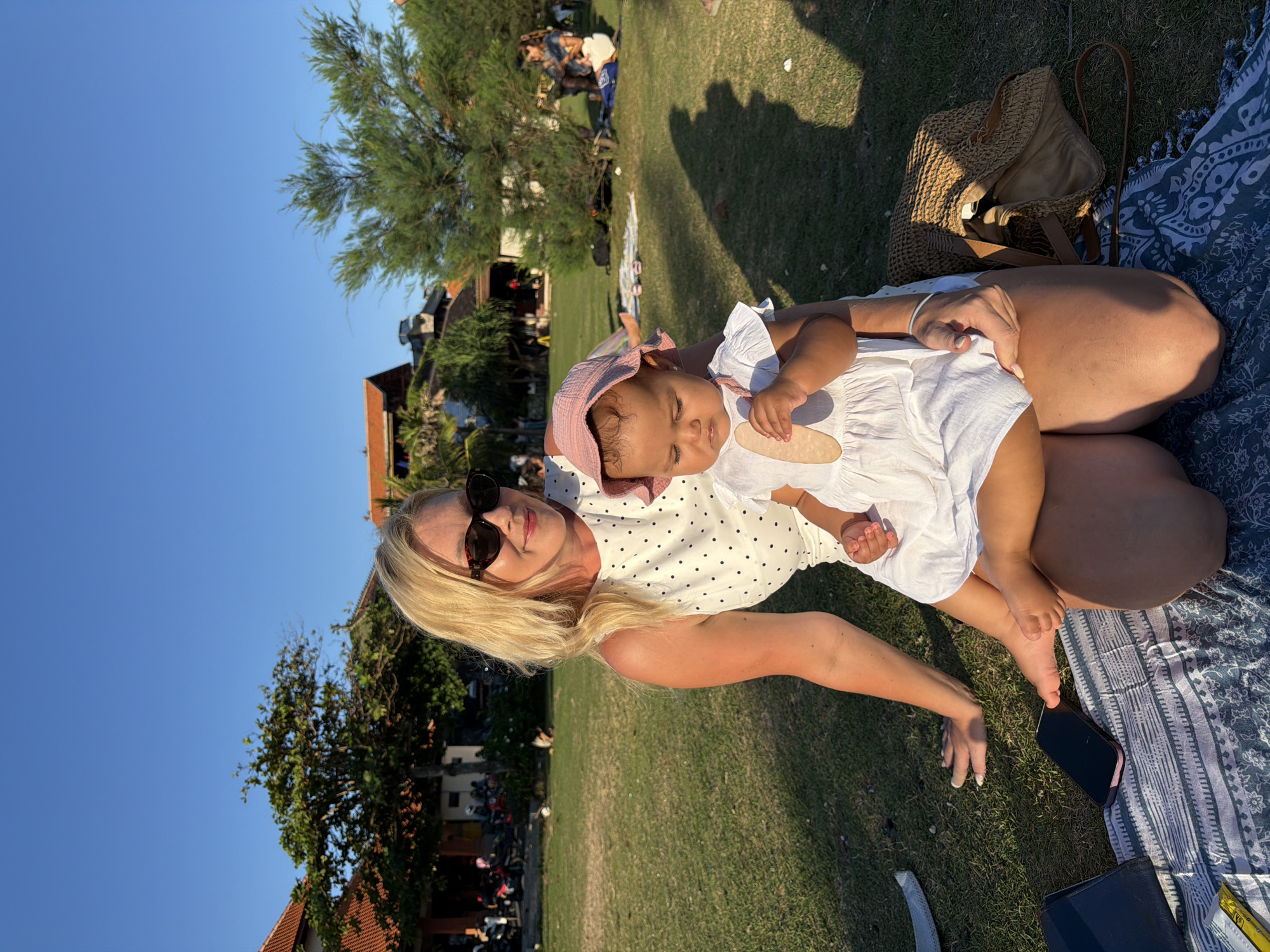 In a park-like setting in Mengwi, Indonesia, a blonde woman wearing sunglasses and a white polka dot dress sits on a picnic blanket on a grassy area. She is holding a baby girl, who is wearing a pink bonnet and a white dress. The baby is looking down at something in her hand. Several trees and buildings with terracotta roofs are visible in the background. The sun is casting long shadows, indicating it is late afternoon or early evening. A woven straw bag is placed next to the woman on the blanket. A smartphone and a book are also on the blanket.