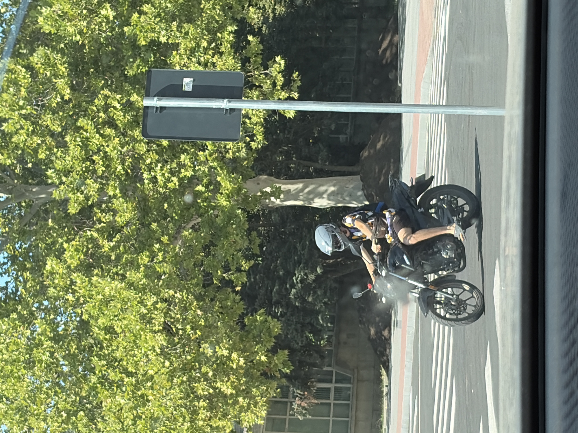 A person wearing a helmet and a patterned shirt rides a black motorcycle on a street with a crosswalk. The person is wearing shorts and sandals. In the background, there are trees, a building, and a traffic sign. The image appears to be taken from inside a vehicle. The location is the city of Criuleni, Moldova.