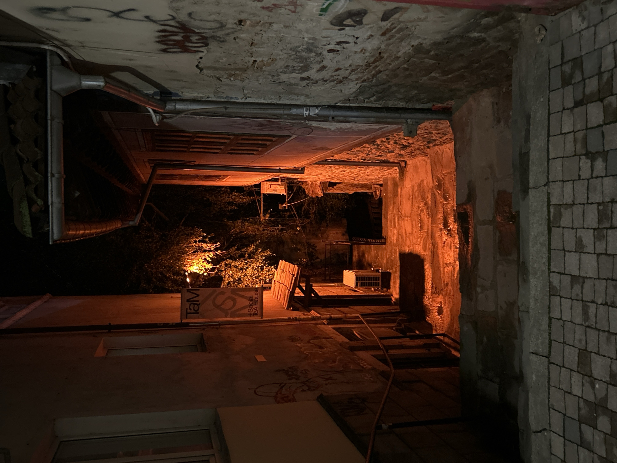 The image depicts an alleyway in what may be Veliko Tarnovo, Bulgaria. The alley is narrow, with aged brick and stone walls. Graffiti is visible on the ceiling. A building with windows and signage, including "Social Tea House", borders one side of the alley. There's a light source casting an orange glow at the far end, where some wooden structures and an air conditioning unit are placed. The scene is dimly lit and appears to be nighttime.