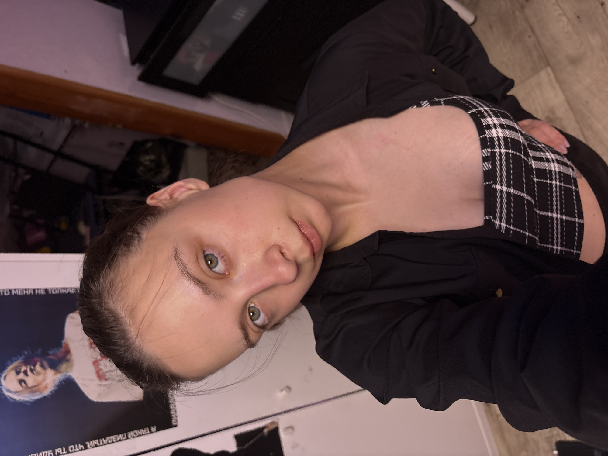 A woman is pictured in a close-up selfie. Her head is tilted to the left, and she is looking directly at the camera. She has fair skin, light green eyes, and dark brown hair pulled back from her face. She is wearing a black button-down shirt over a black and white checkered top. The background is cluttered and includes a wooden floor, a dark-colored object (possibly furniture), and a white surface with some writing in Russian. There are no details to determine if this picture was taken in Прикамский, Russia.