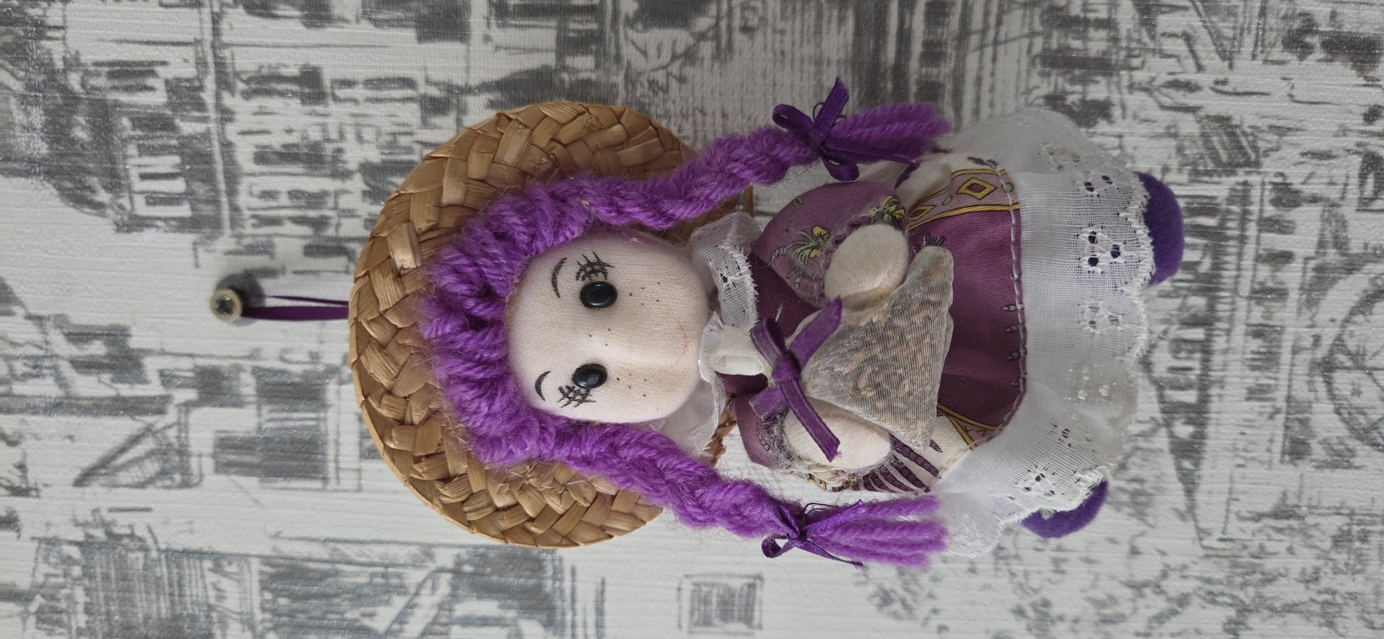 The image displays a handmade fabric doll, vertically oriented but depicting the doll standing or hanging upright. The doll has a light-colored, soft fabric face with black button eyes, embroidered black eyebrows, and a simple, slightly smiling mouth. Pink blush is visible on its cheeks. Its hair is bright purple yarn, styled into braided pigtails secured with small purple ribbons. A woven straw hat sits atop its head.
The doll wears a purple fabric dress with a patterned border featuring gold and white designs. A white lace apron or overlay with delicate eyelet embroidery covers part of the dress. In its hands, the doll holds two small, light-colored fabric bundles, each tied with a purple ribbon. These bundles appear to contain granular material, possibly dried lavender or similar aromatic contents, suggesting they are sachets. Purple fabric is also visible at the doll's lower extremities, likely part of its attire or footwear.
The doll is suspended from a wall or surface, indicated by a visible silver screw with a thin purple ribbon attached, positioned above the doll's head. The background is a light-colored surface adorned with a subtle, repeating grey pattern resembling architectural outlines or stylized city buildings, suggesting wallpaper or a printed surface. The scene is static, depicting the doll as a decorative item. No text is visible in the image.
