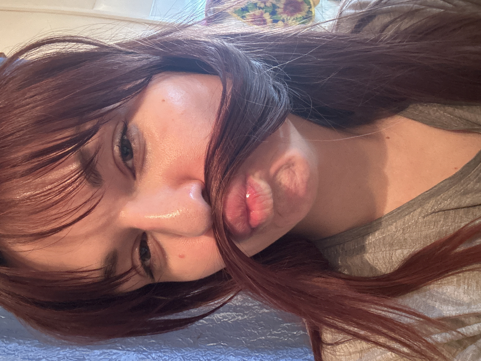 A woman with reddish-brown hair is lying on a blue surface, possibly a bed, with her head tilted towards the viewer. She is wearing a gray t-shirt. Her hair is partially covering her face. She is making a pucker face. The image appears to be taken indoors. There is no information to suggest the location is in the city of Фоки, Russia.