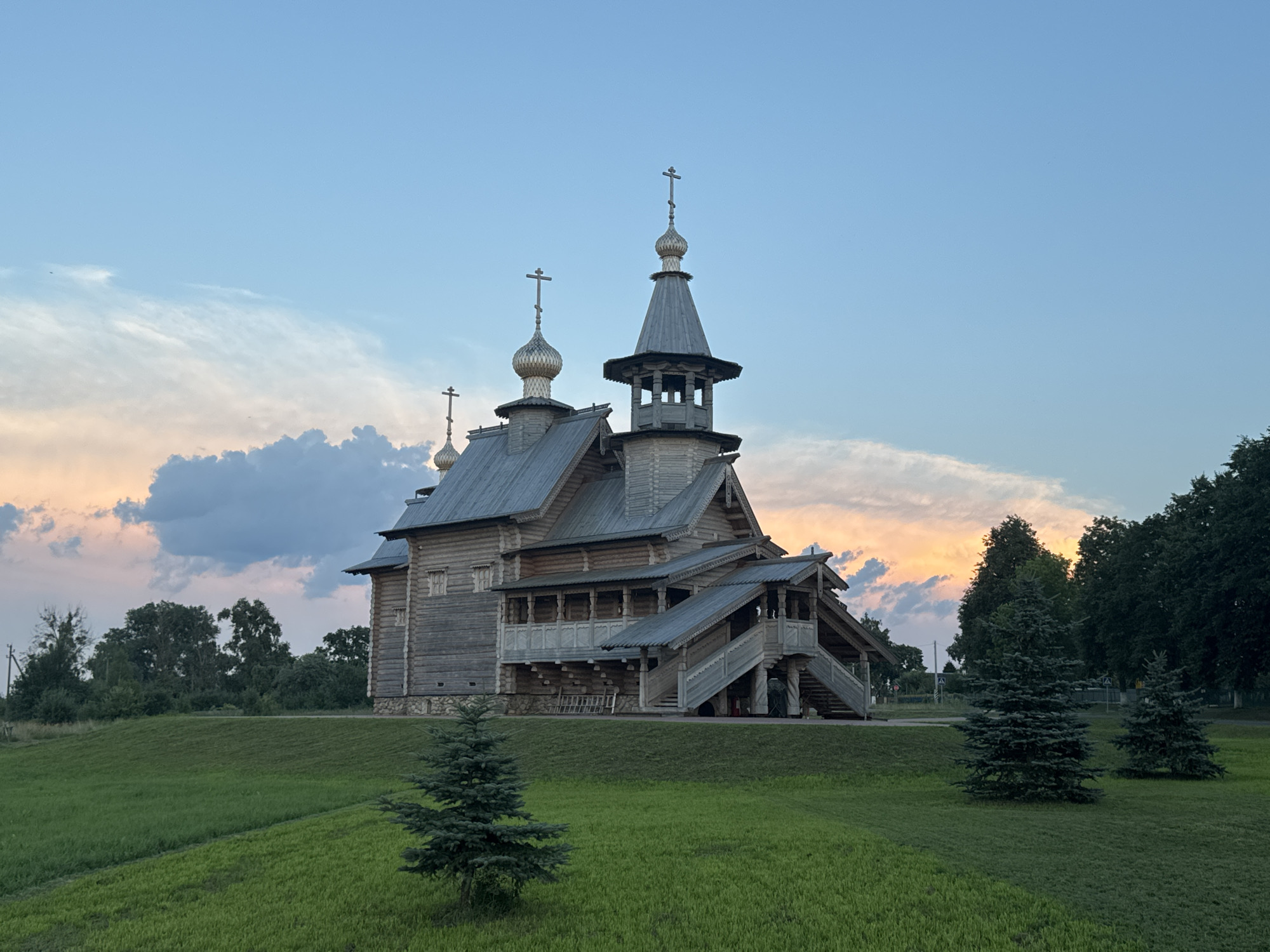An exterior view of a traditional wooden Orthodox church with multiple onion domes and crosses is captured under a twilight sky. The church, constructed from logs, features intricate wooden detailing, including a covered walkway and a prominent staircase leading to an elevated entrance. The building is situated on a grassy hill, with several young evergreen trees in the foreground. In the background, a line of mature trees forms a dense forest, and a faint sunset can be seen through the clouds. The sky is a gradient of blue, transitioning to pale orange and pink hues near the horizon. There are no people visible in the scene. The location is identified as the village of Neman, in the Neman Rural Council, Belarus.