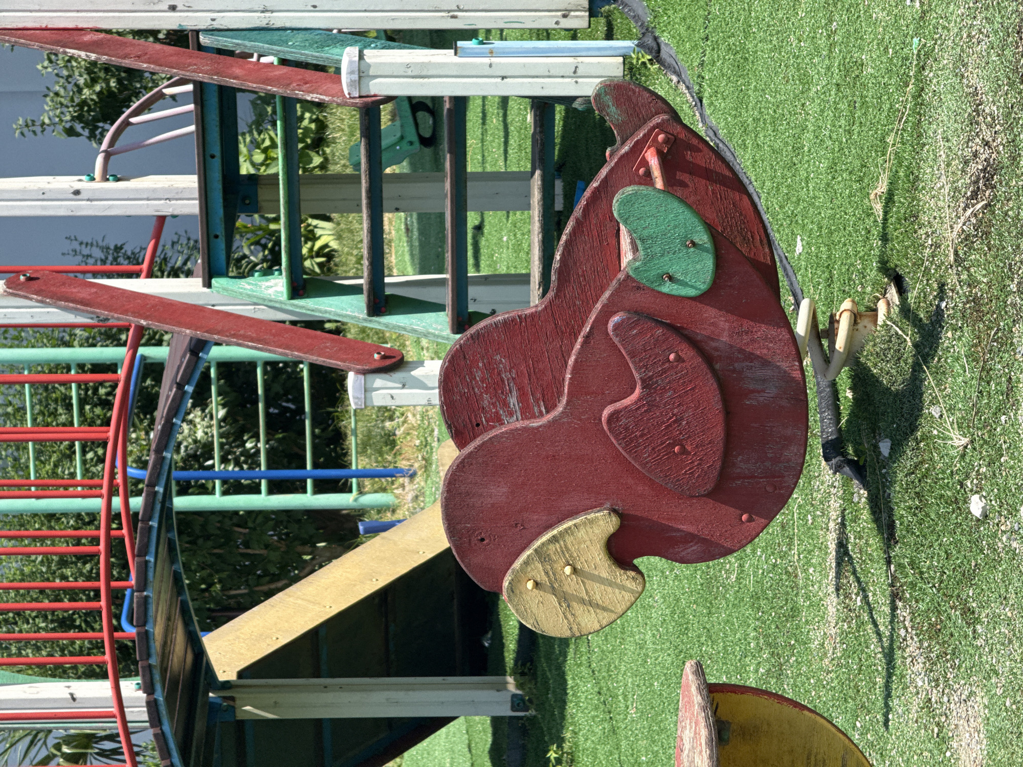 The image shows a red wooden parrot-shaped spring rider on a green artificial turf playground. The rider is partially obscured by other playground equipment, including a yellow slide and a set of stairs with green and blue rungs. In the background, there are red and green metal bars and posts of a larger playground structure. The environment appears to be outdoors in a sunny, urban setting, likely a park or schoolyard. No people are visible in the image. The ground is covered in artificial grass with scattered small stones. The playground equipment appears to be made of wood and metal, showing some signs of wear and tear. The overall scene depicts a children's play area in a city.