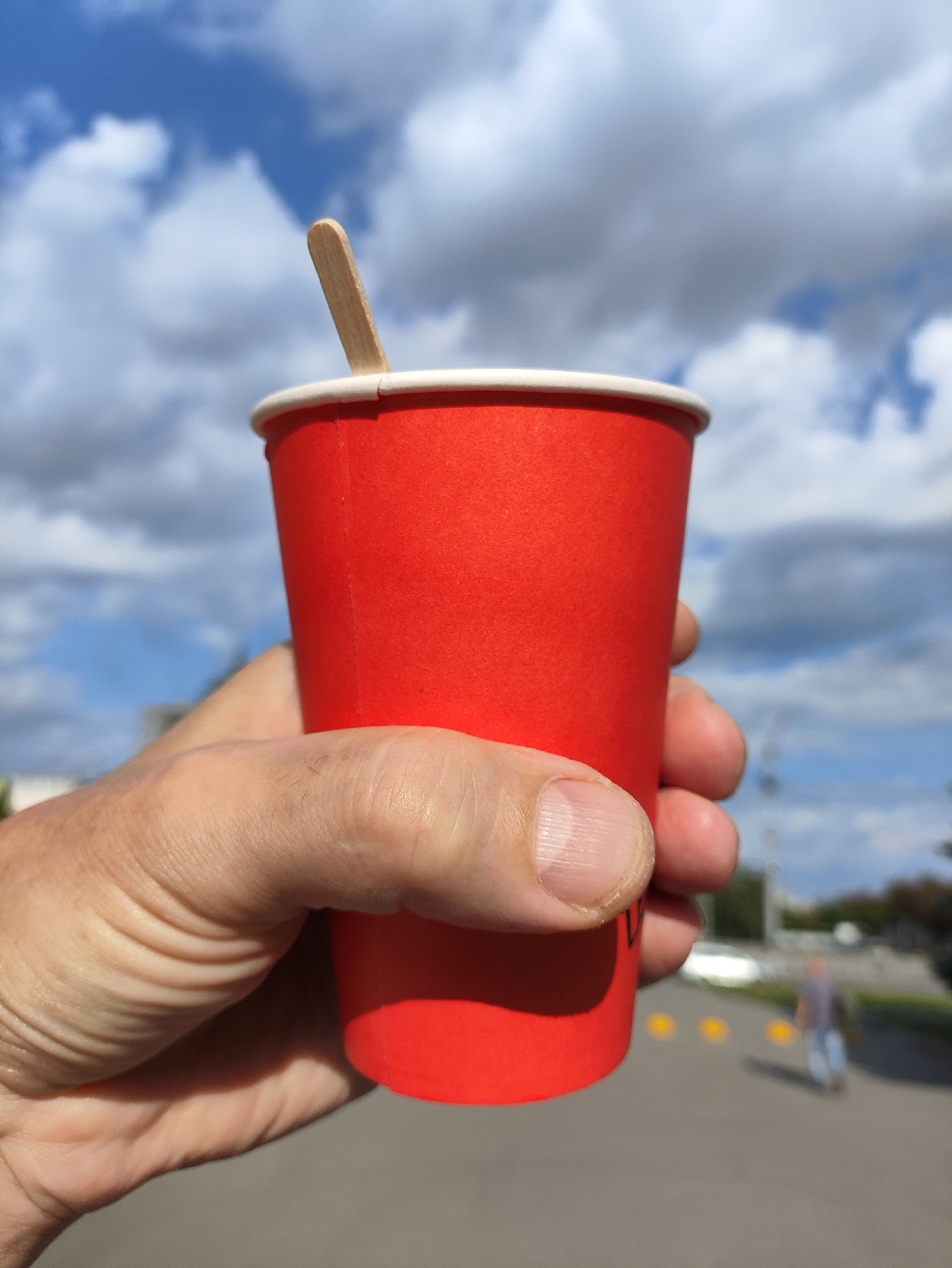 A person's hand holds a bright red paper cup with a white rim. A wooden stirrer protrudes from the top of the cup. The cup is held in the foreground, with a blurred background of a cloudy blue sky and a paved surface with some yellow markers. A blurry figure of a person walking away is visible in the distance.