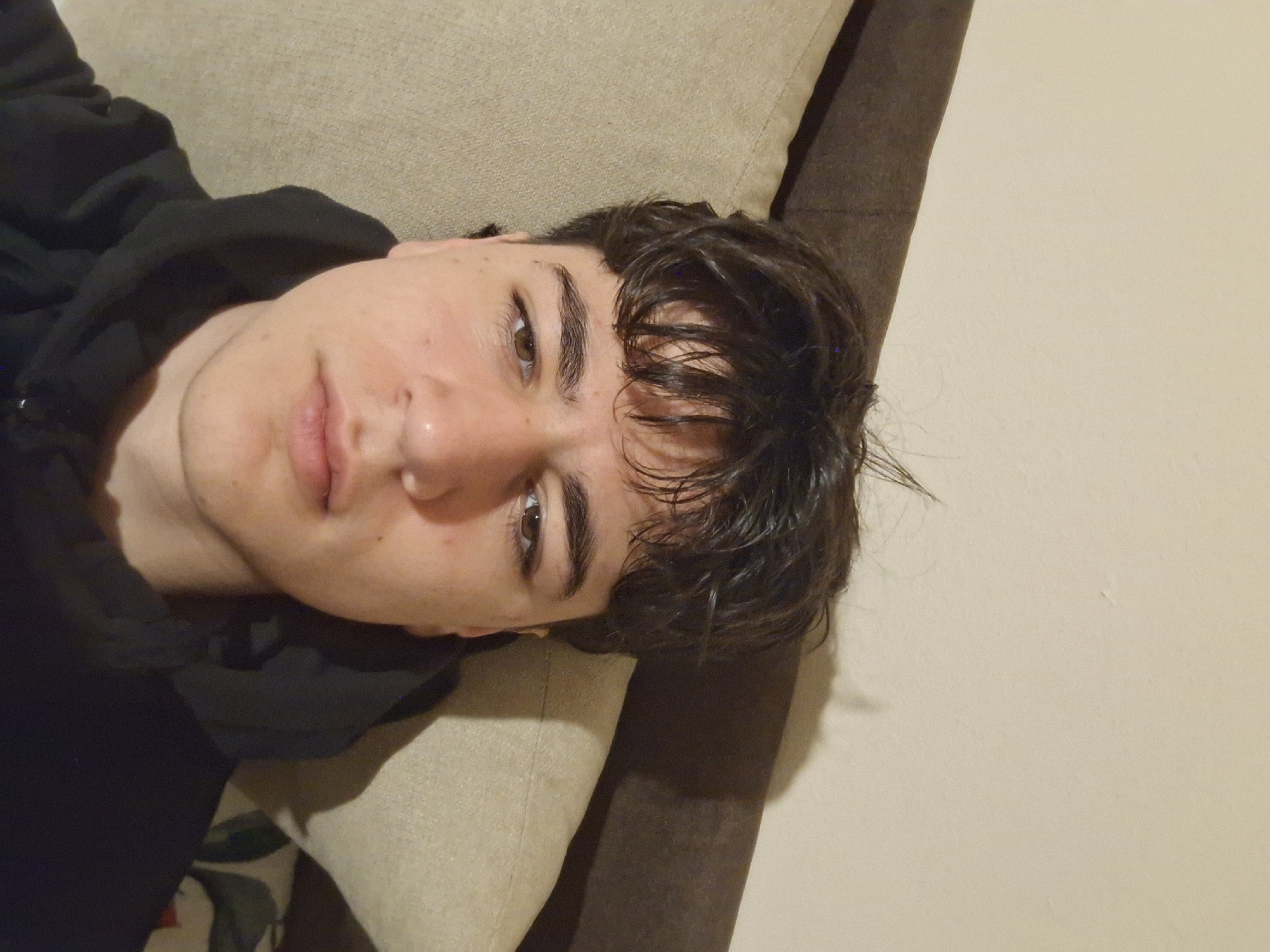 A young man is lying on a beige and brown couch. He is wearing a black hoodie. His hair is dark and appears to be wet or styled with product. The background is a plain, light-colored wall. The location (Burgas, Bulgaria) cannot be verified from the visual information provided.