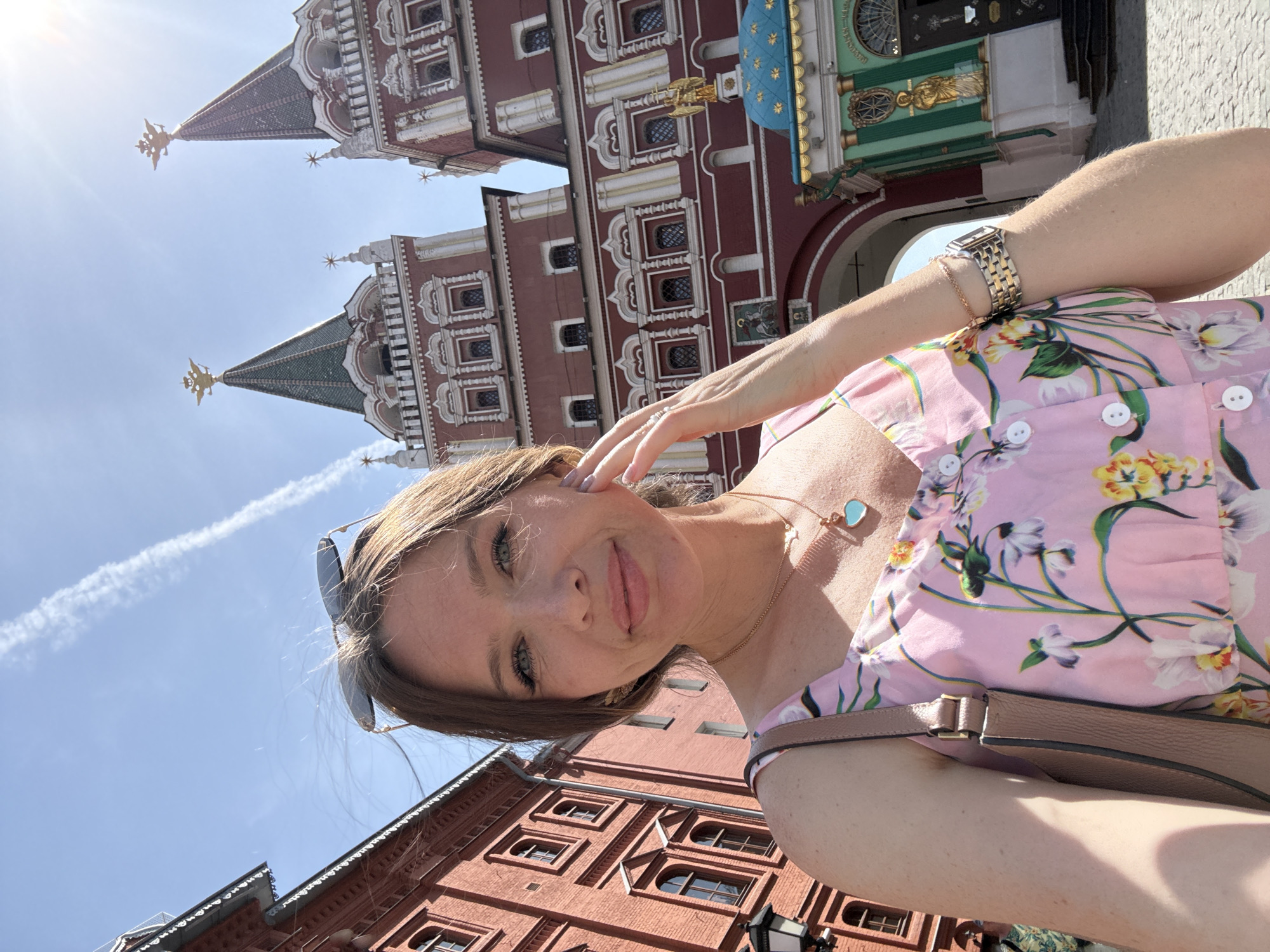 A woman is taking a selfie in Moscow, Russia with the Resurrection Gate and parts of the State Historical Museum visible behind her. She is wearing a pink floral dress, a wristwatch, and a necklace. The background includes red brick buildings and a clear blue sky. The sun is shining brightly, and there is a contrail visible in the sky.